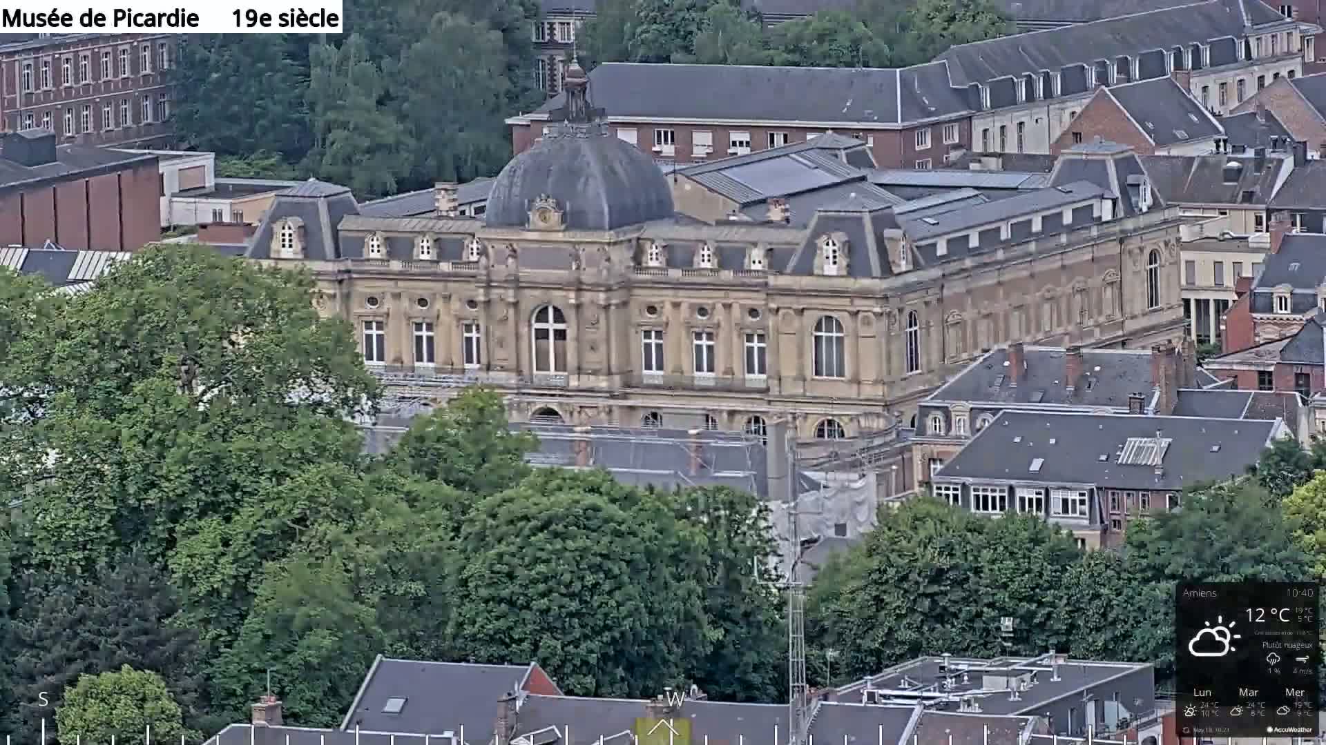 A large, light-brown stone building with a dome sits amidst green trees and other buildings on a partly cloudy day.