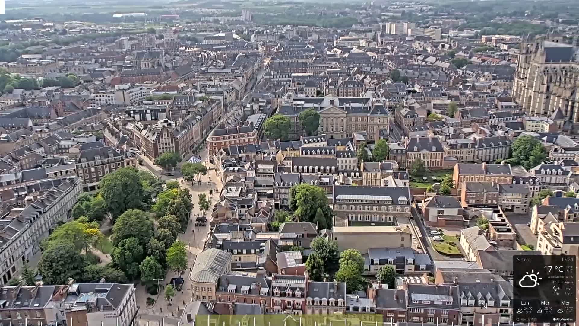 A high-angle, long shot of a European city on a partly cloudy day, showing many closely-packed buildings and a central square with green space.