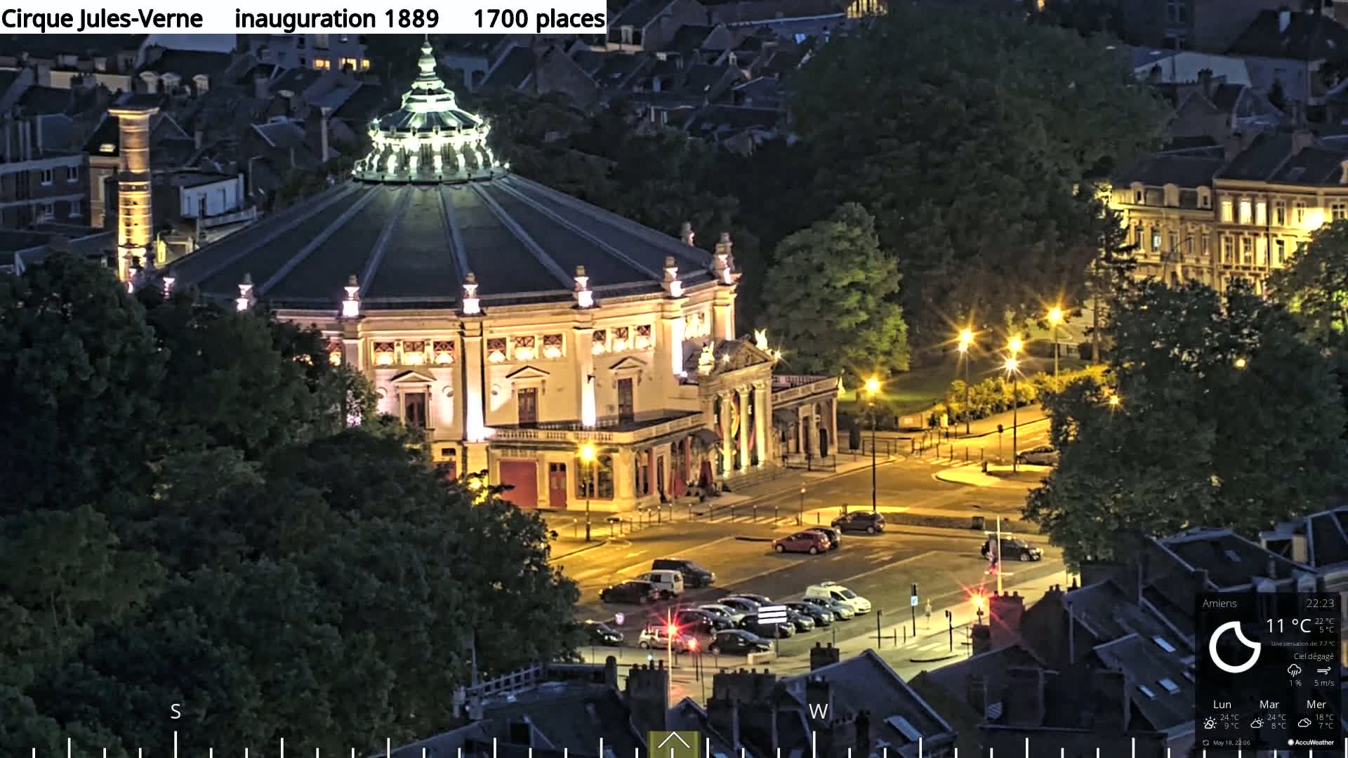A large, round, illuminated building sits in a city square at night, surrounded by parked cars and dark trees under a clear sky.