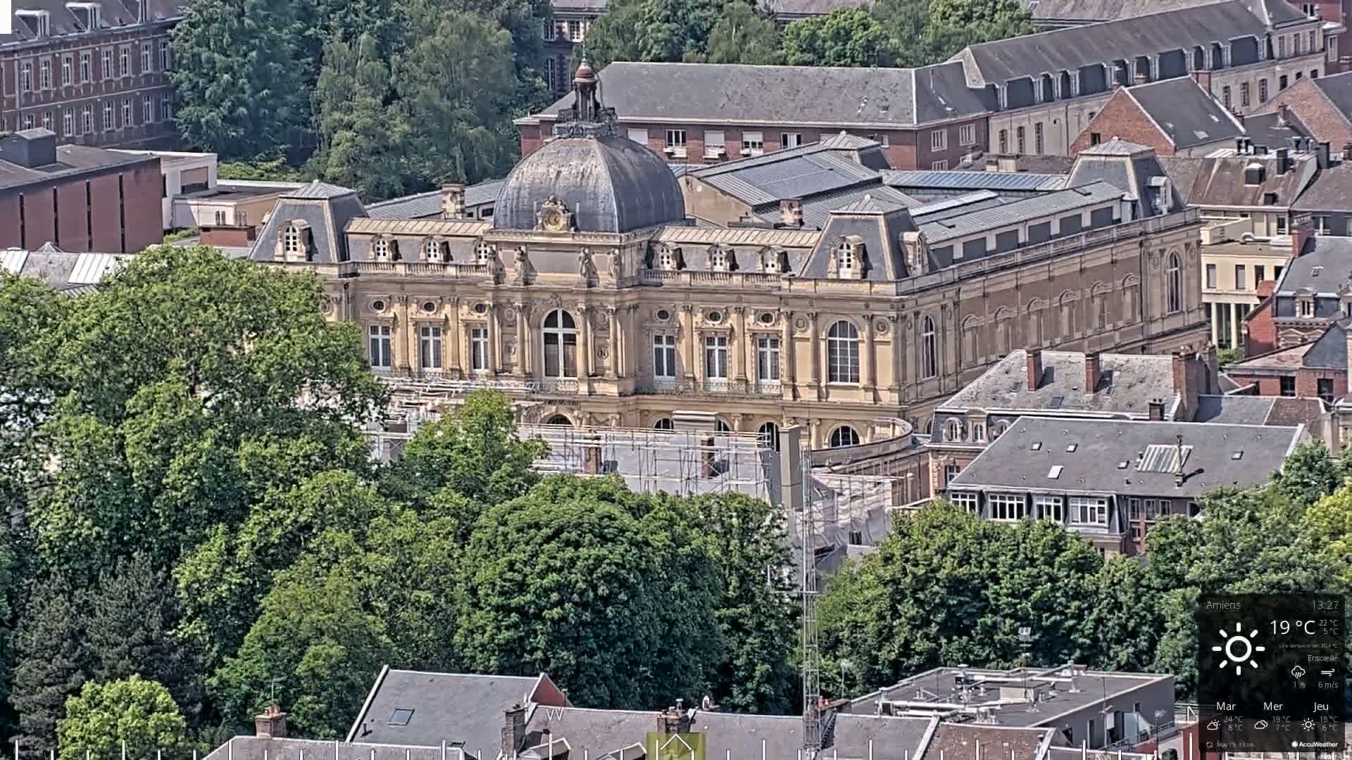 An aerial view shows a large, tan building with a dome, surrounded by lush green trees and other buildings under sunny skies.