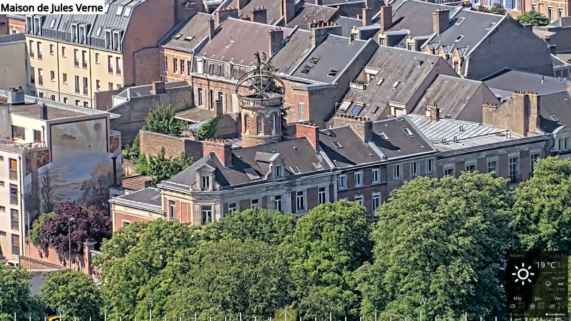 An aerial view shows a large building with a dark grey roof, surrounded by other buildings and lush green trees on a sunny day.
