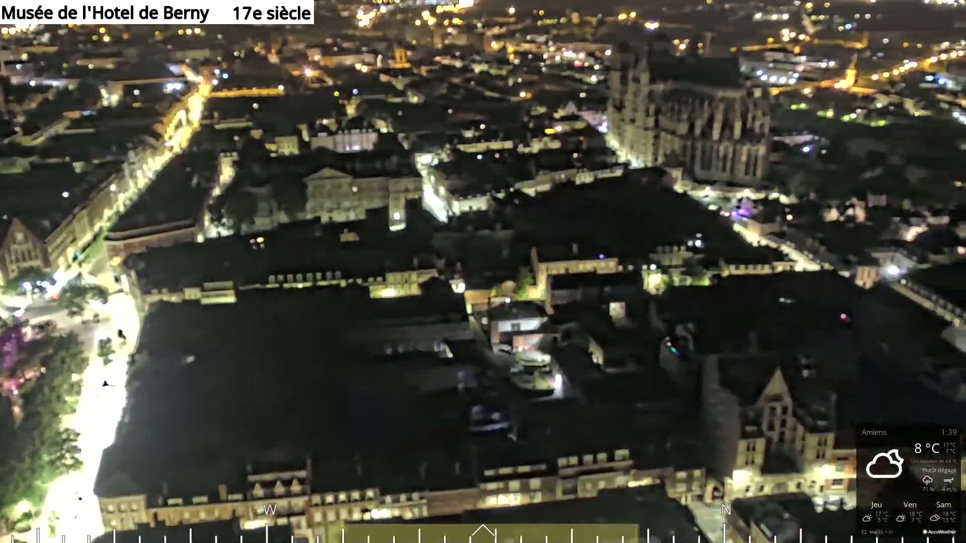 A nighttime aerial view of a city with illuminated streets and buildings, including a large cathedral, under partly clear skies.