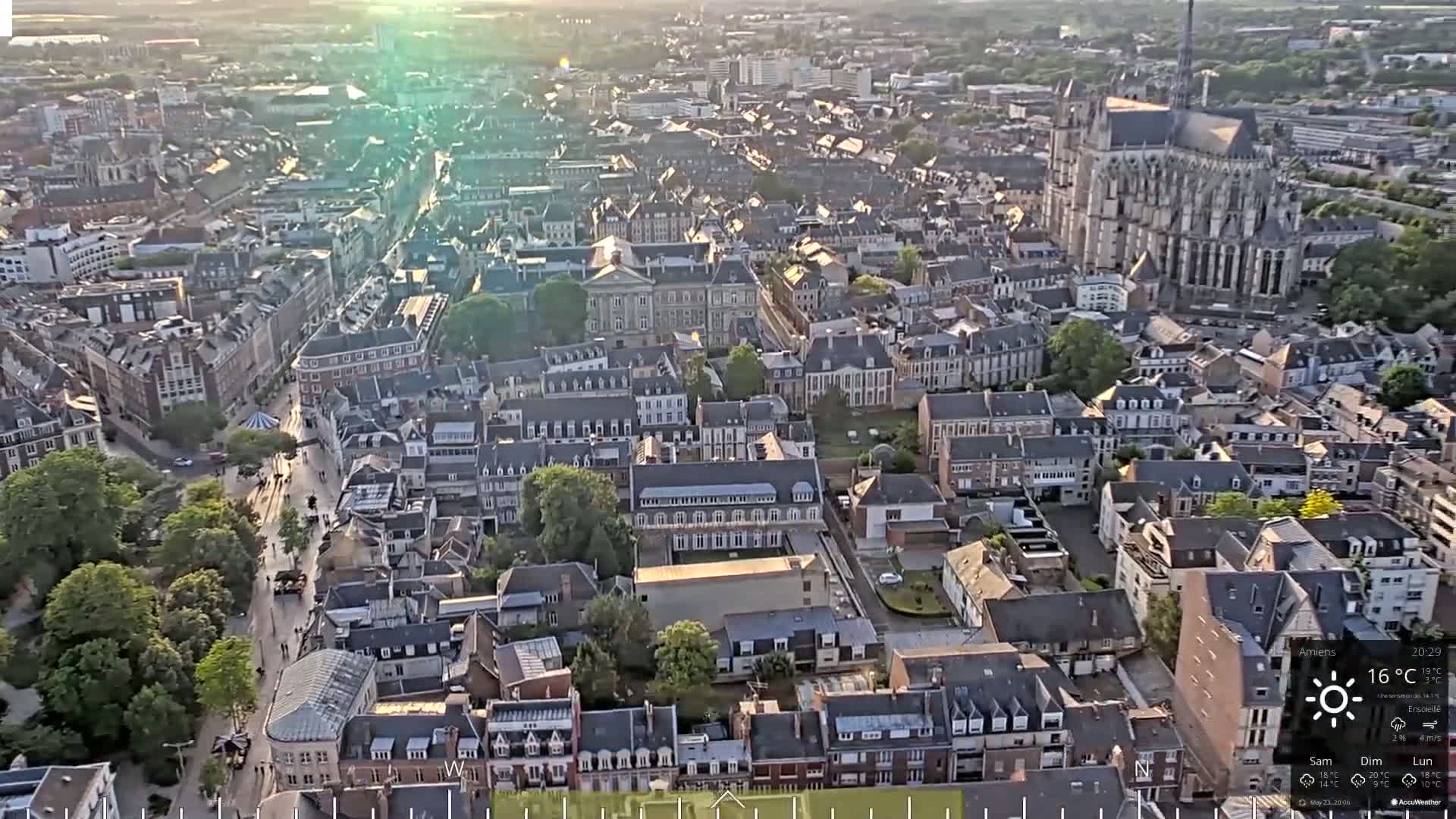 An aerial view of a European city on a sunny day, showing a dense collection of buildings, including a large cathedral, and a street running through the city center.
