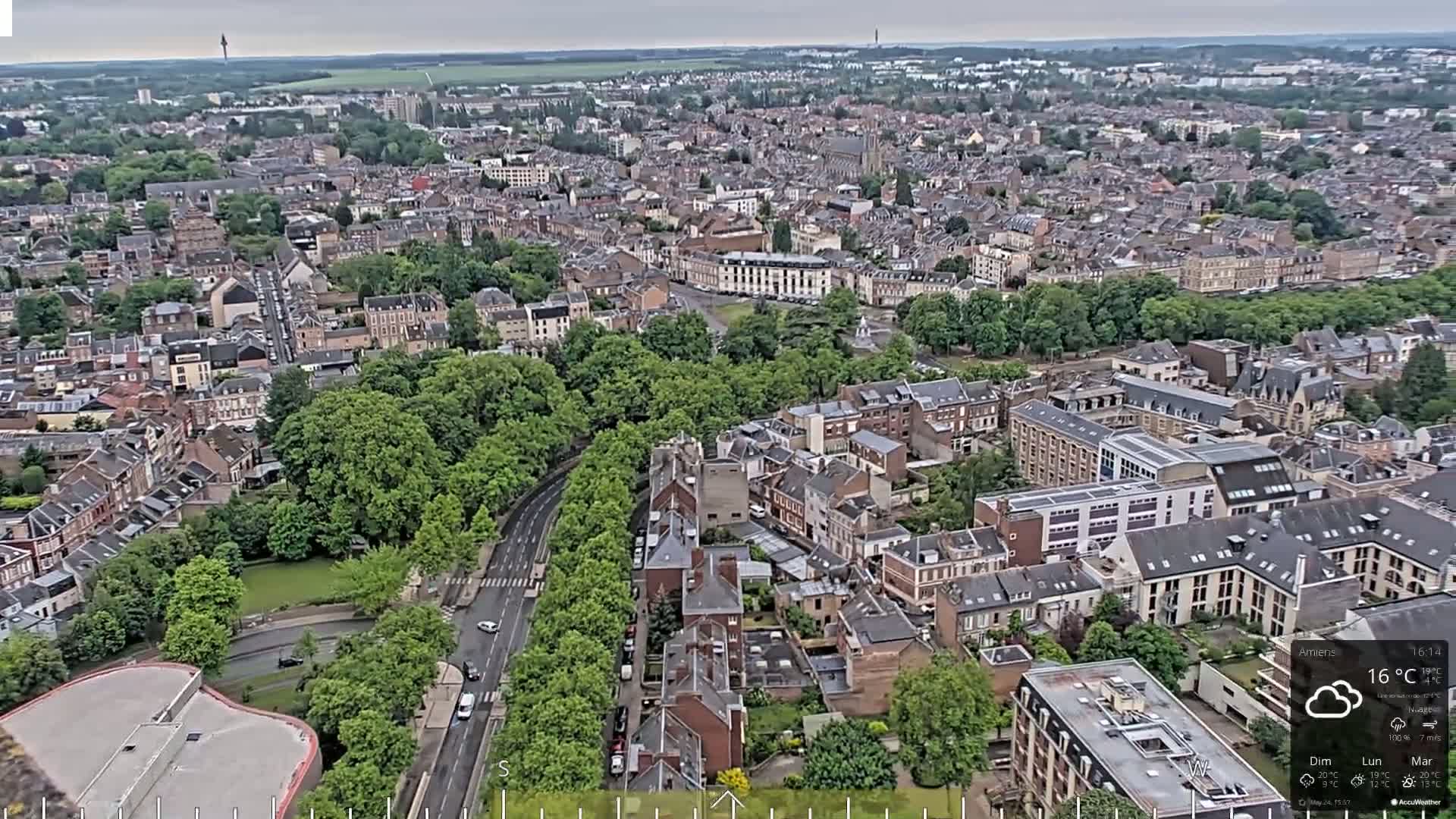 An aerial view shows a city on an overcast day, with numerous buildings, green spaces, and roads visible from a high vantage point.