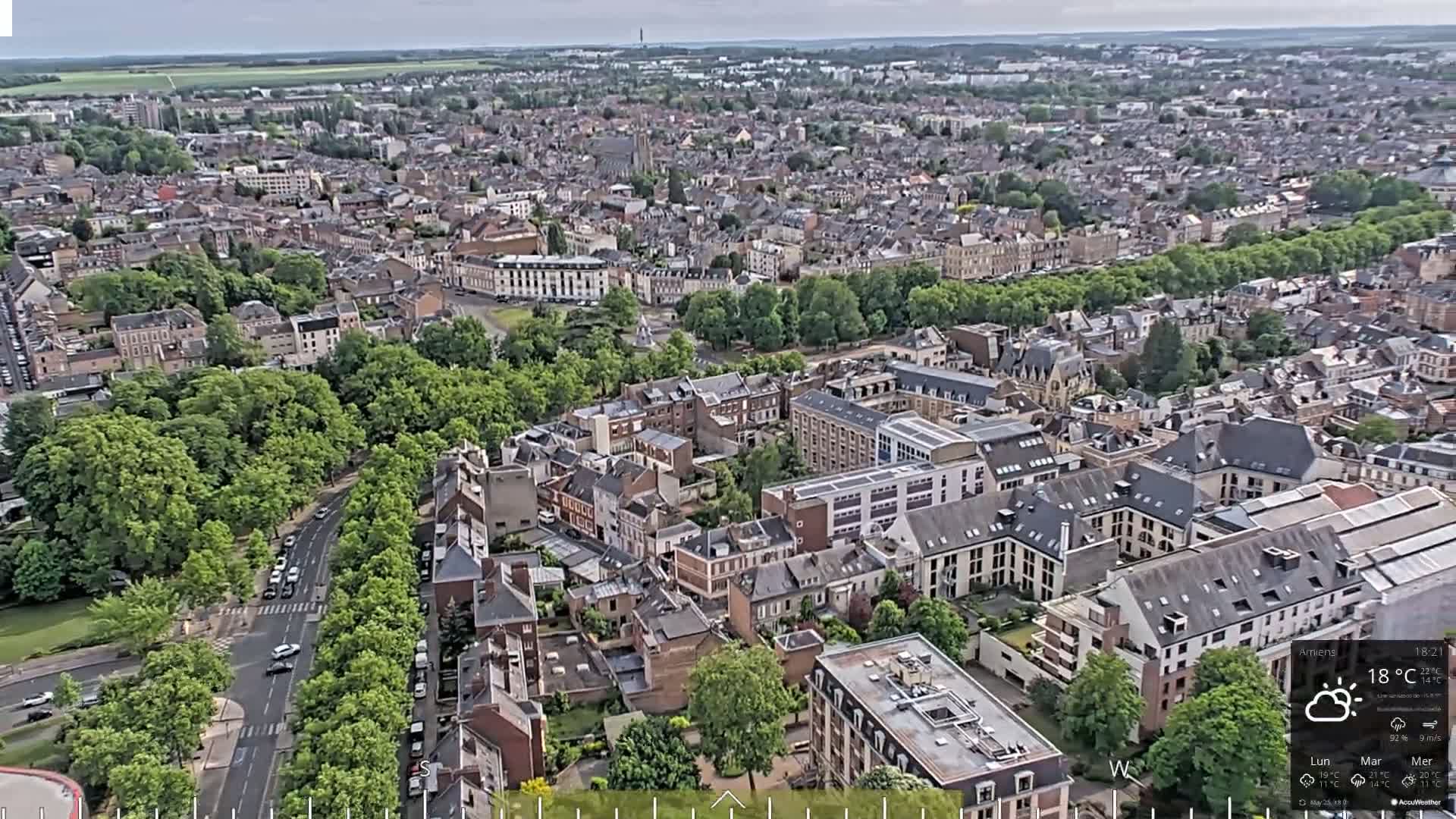 An aerial view of a city on an overcast day shows many tightly packed buildings surrounded by green trees and roads.
