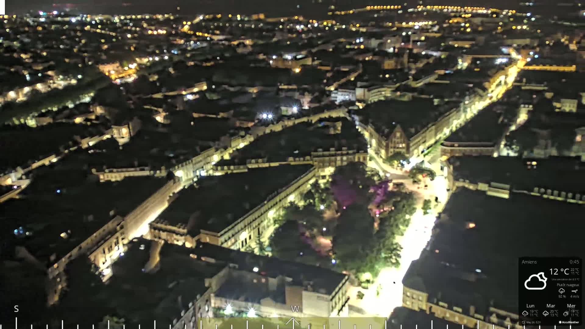 An aerial nighttime view of a city with illuminated streets and buildings under partly cloudy conditions.