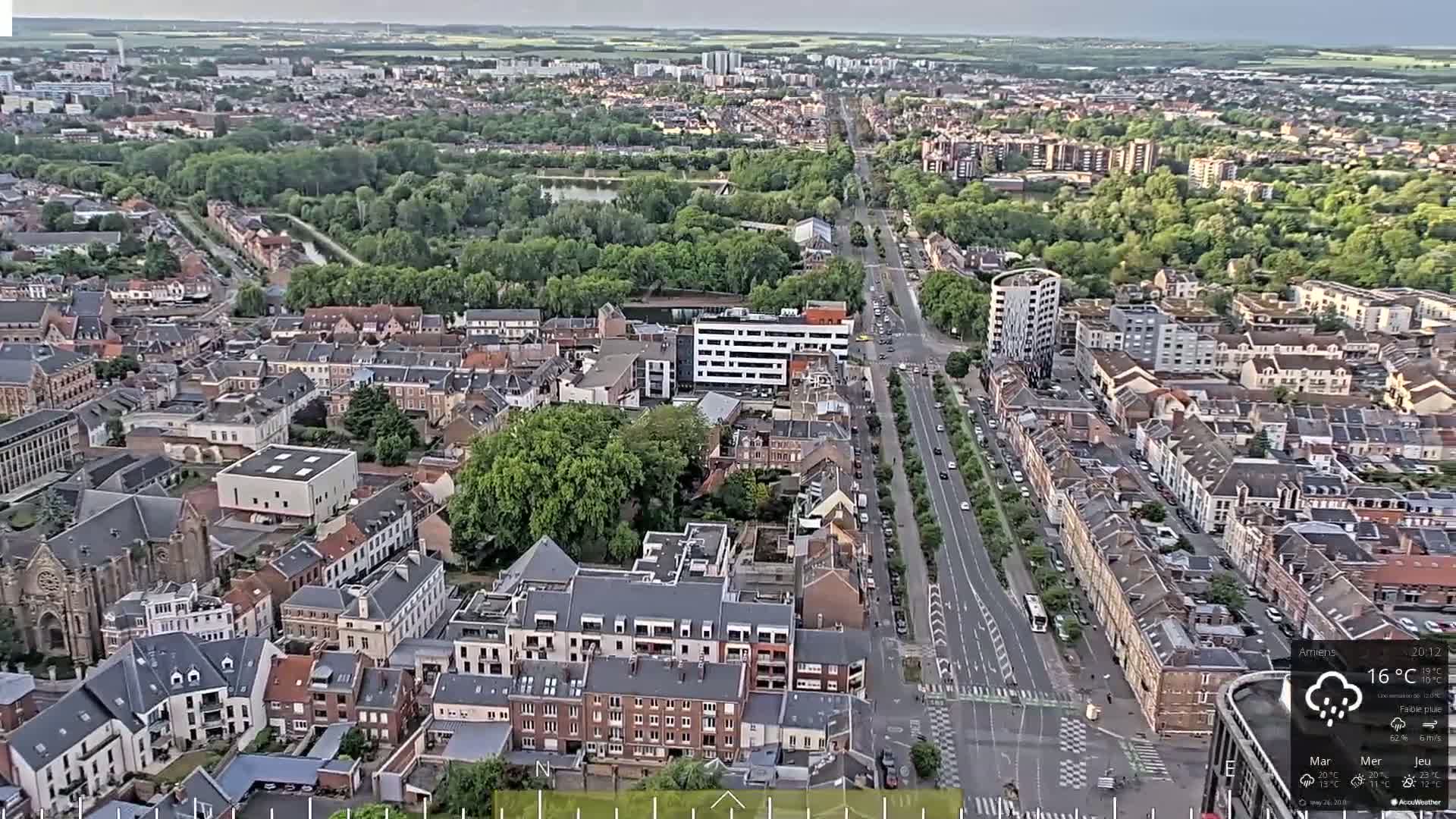 An aerial view of a city on a partly cloudy day, showing a long road lined with buildings and trees, and a wider expanse of urban and suburban areas in the background.