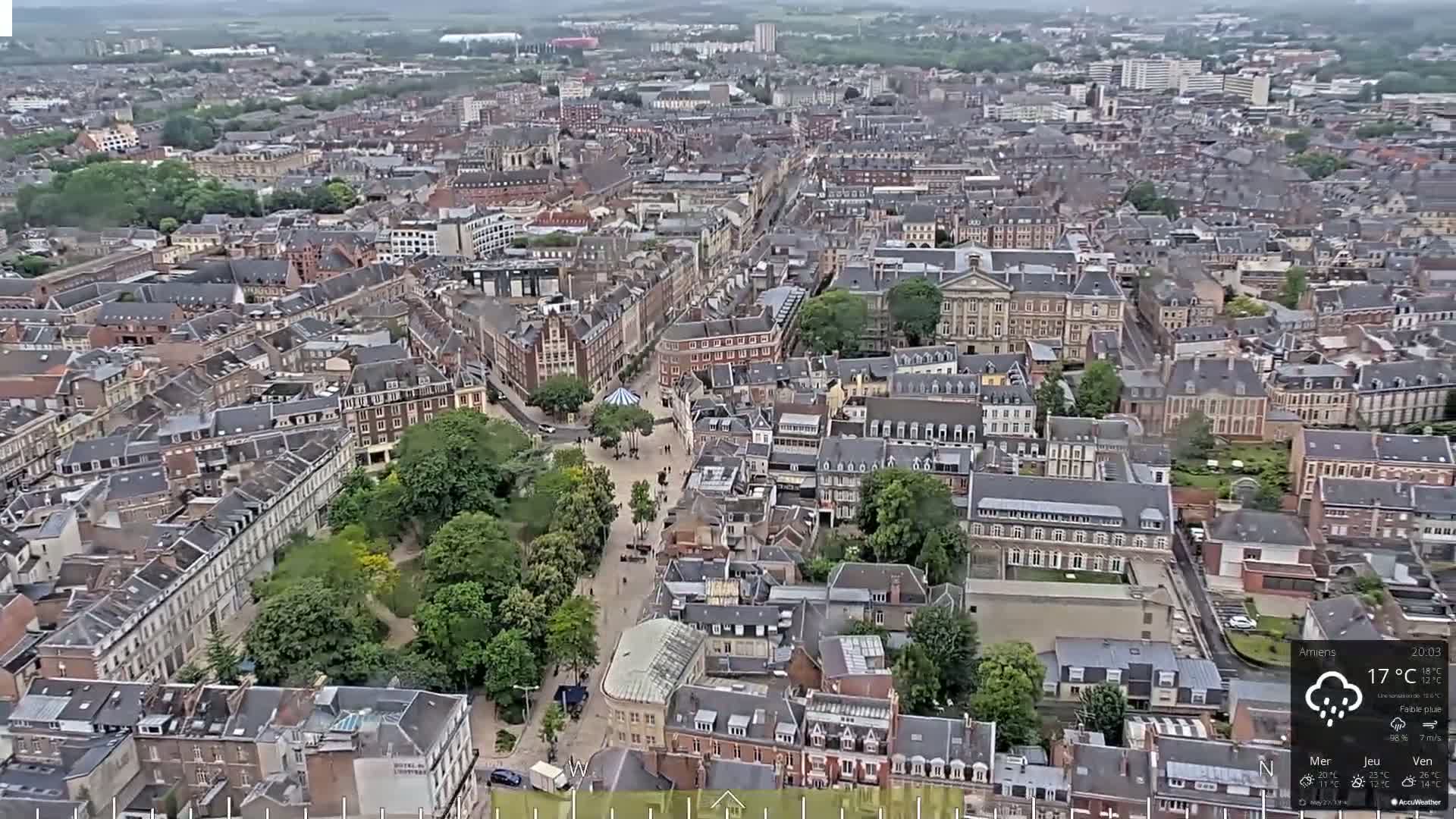 An aerial view of a European city on a cloudy day with light rain shows a dense collection of buildings surrounding a park-like square.