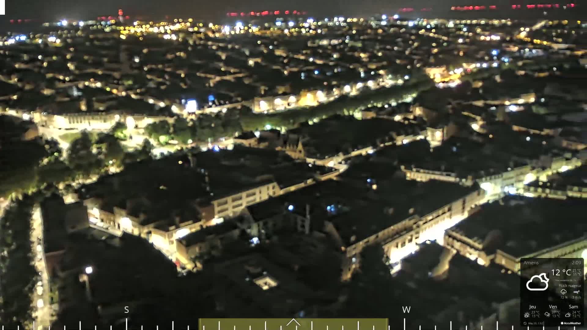 A nighttime aerial view of a city with dimly lit buildings and streets, under partly cloudy skies.