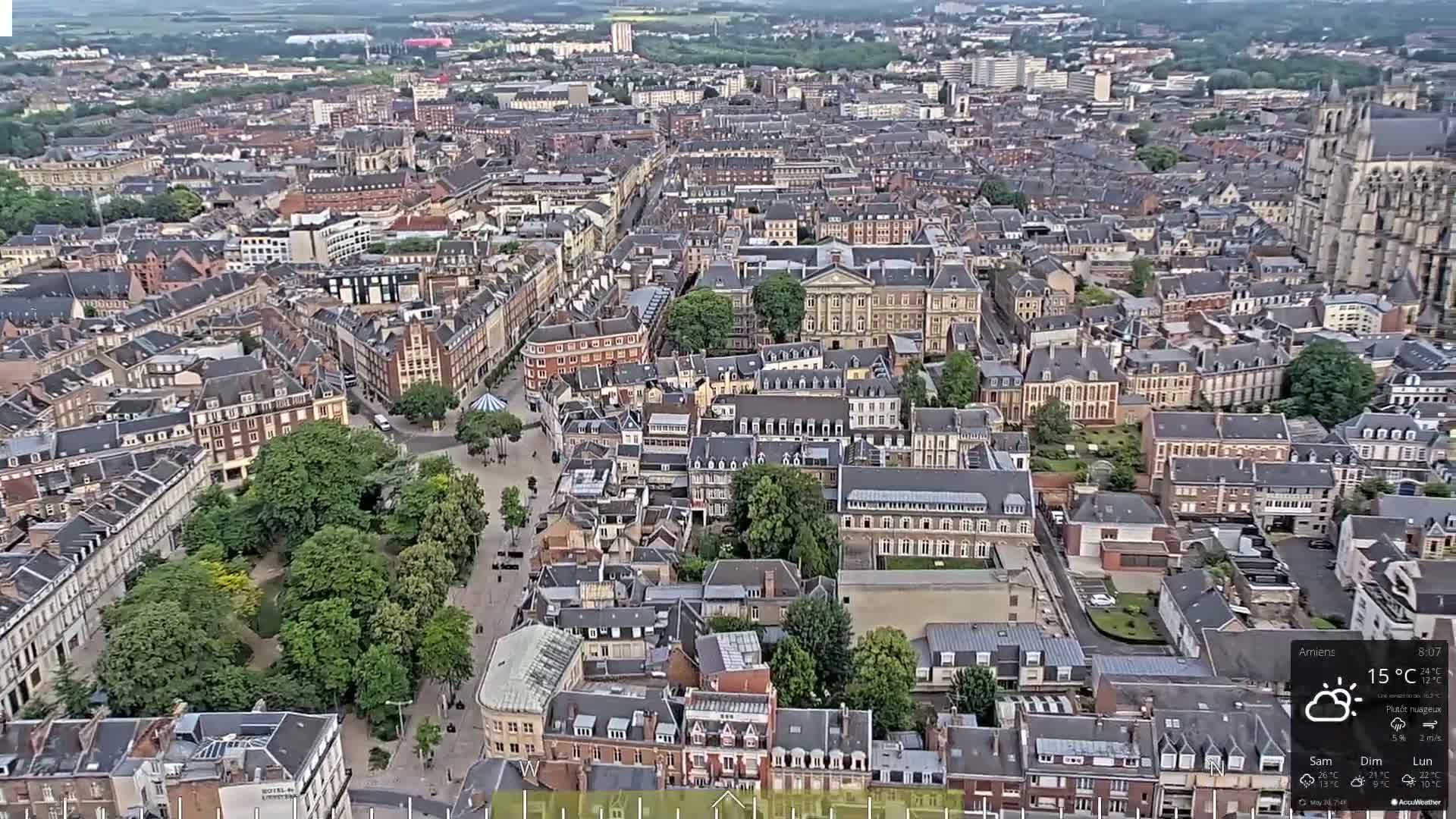 An aerial view of a European city on a partly cloudy day shows a dense collection of buildings surrounding a large, tree-filled square.