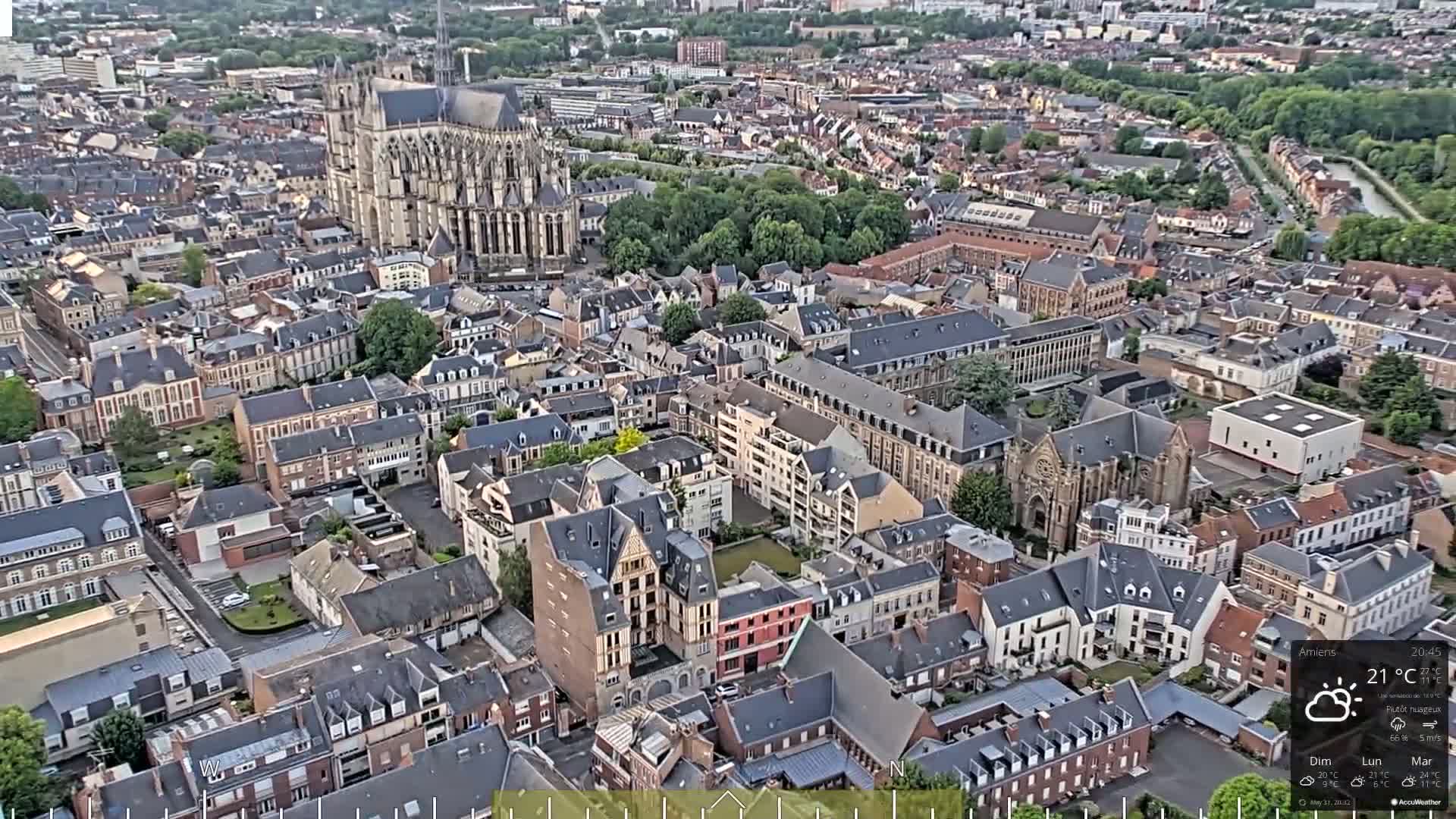 An aerial view shows a European city with a large cathedral and numerous closely packed buildings under partly cloudy skies.