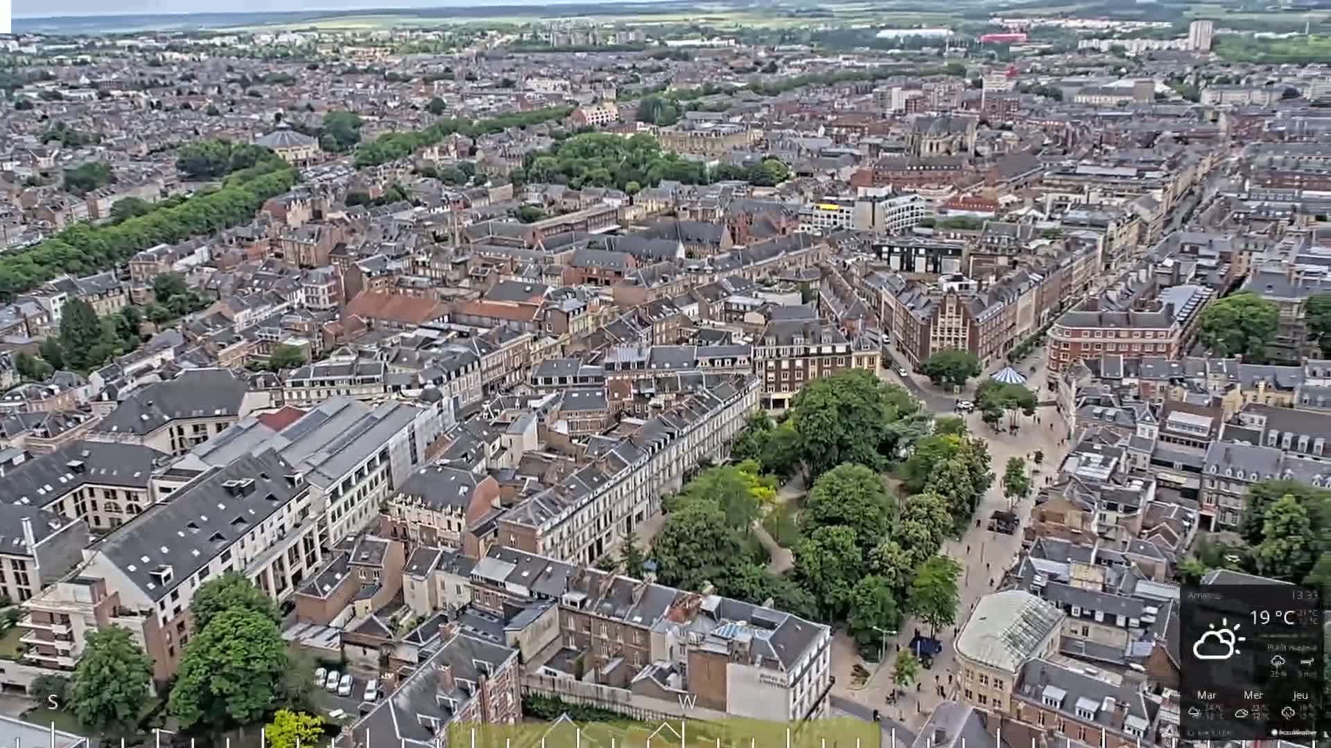 An aerial view of a dense city on a partly cloudy day, showing many closely-packed buildings and a central green space.