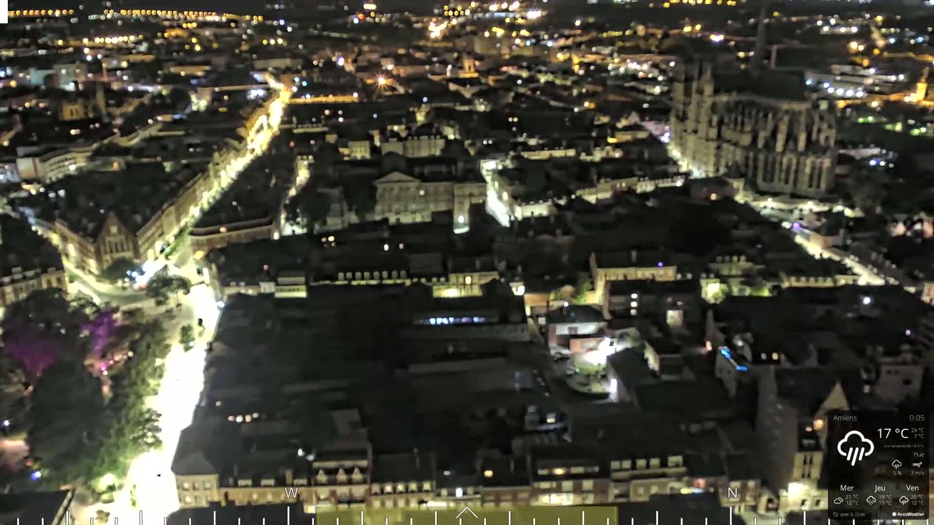 A nighttime aerial view of a city with illuminated streets and buildings, including a large cathedral, under a partly cloudy sky.