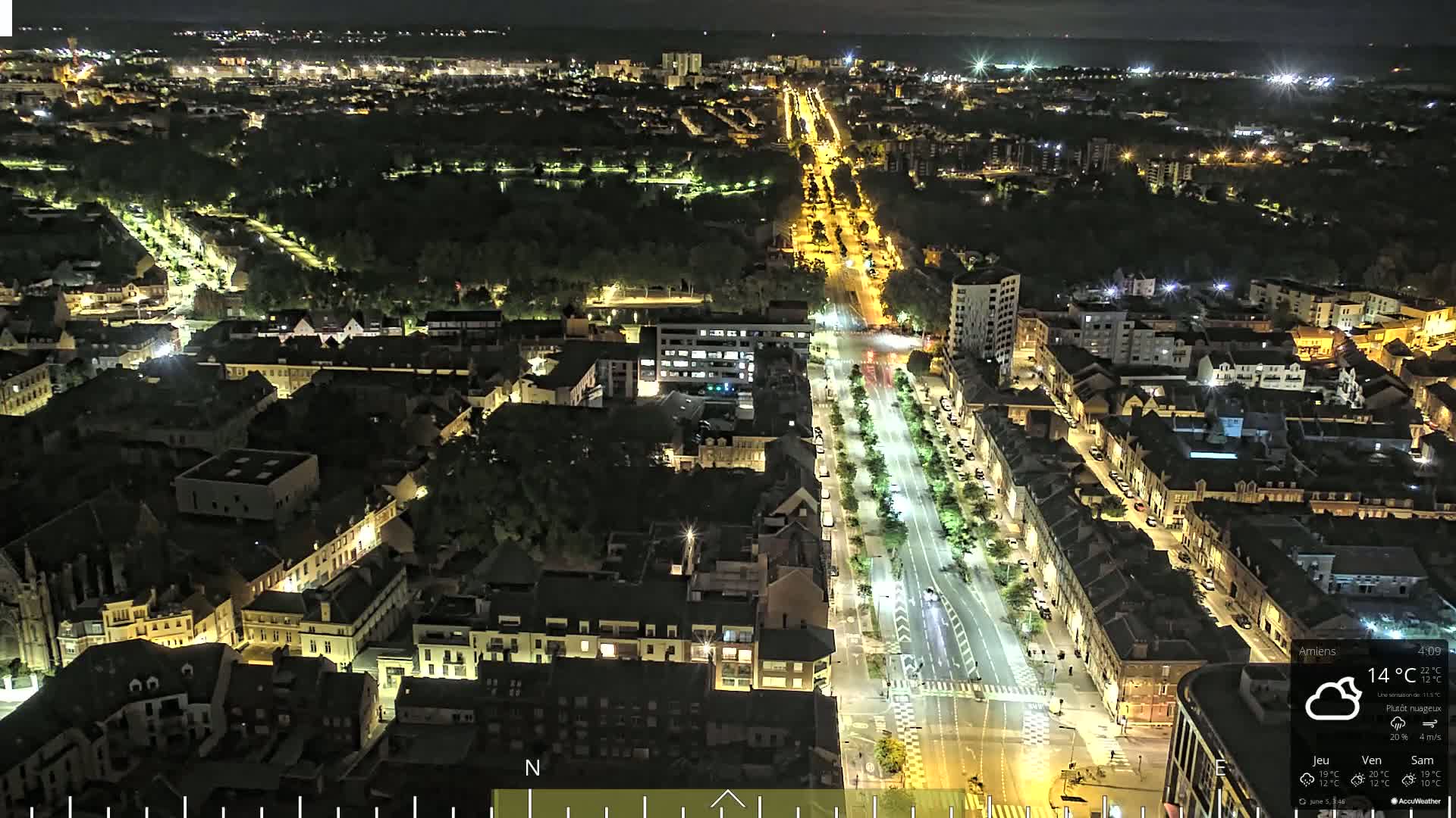 A nighttime aerial view of a city with illuminated streets and buildings, under partly cloudy skies.
