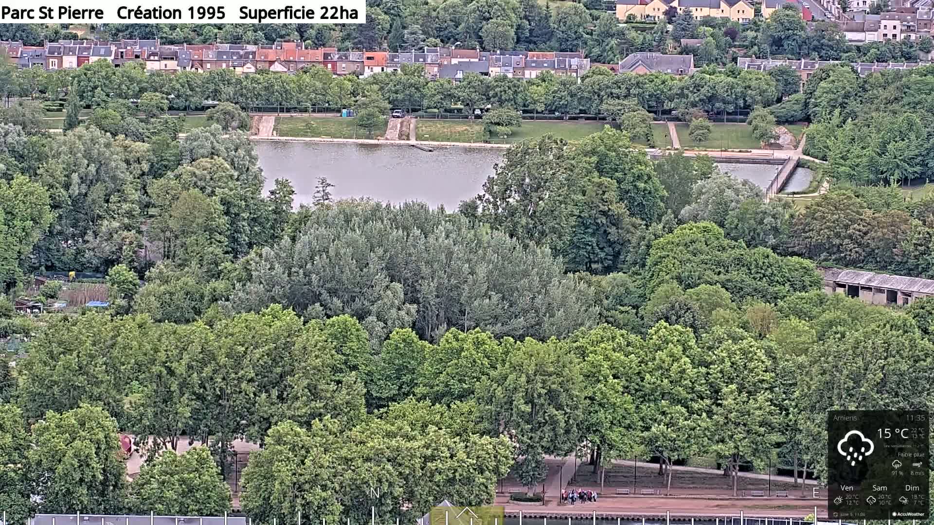 A park with a pond is surrounded by lush green trees and overlooks a residential area under lightly overcast skies with a light rain.