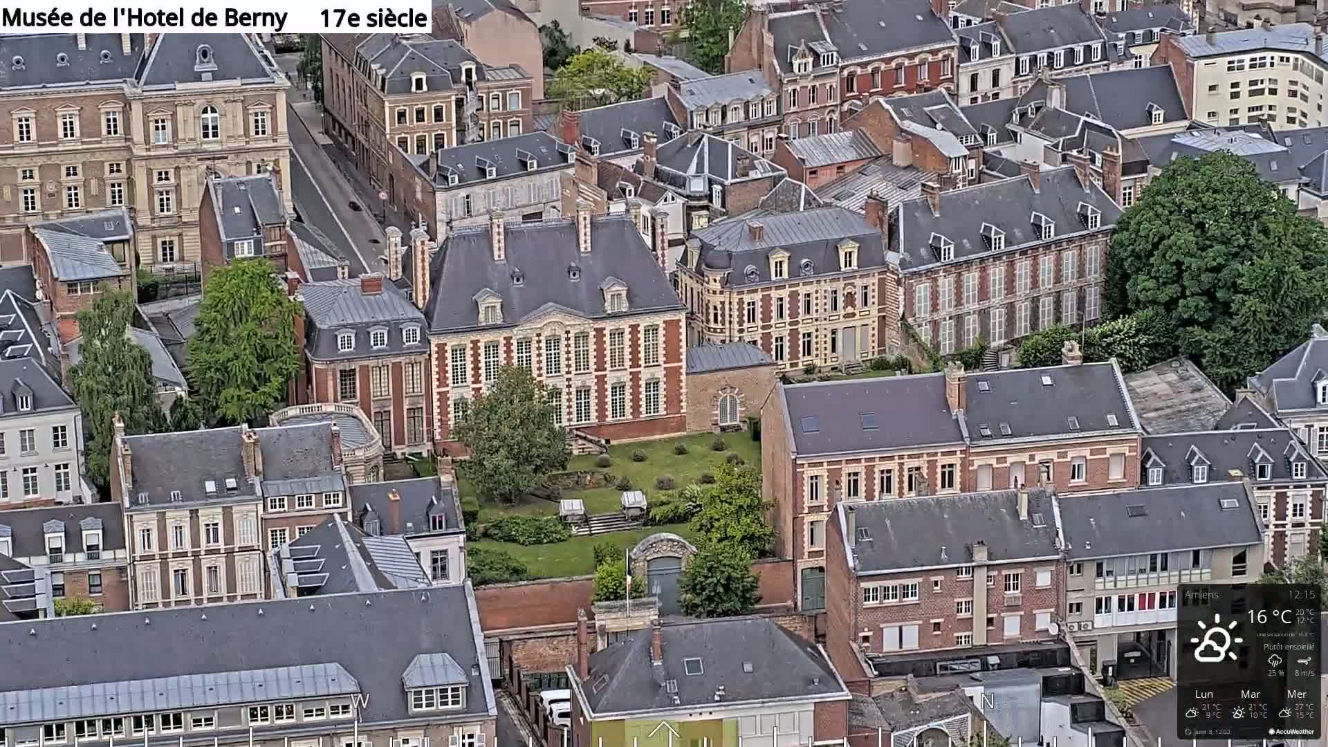 An aerial view shows a large, rectangular building with a garden in the center of a densely packed city under partly sunny skies.