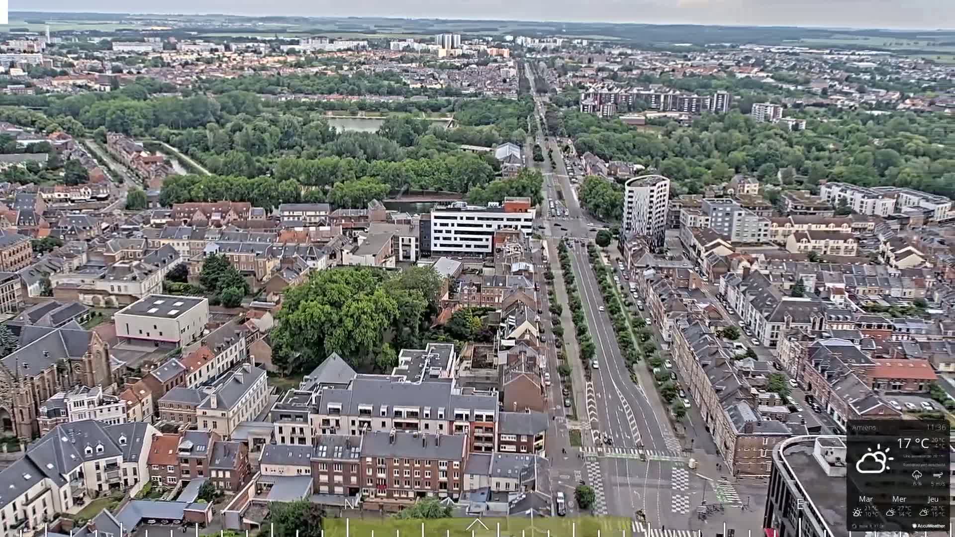 An aerial view of a city on an overcast day, showing a long, tree-lined road extending through a dense residential area.