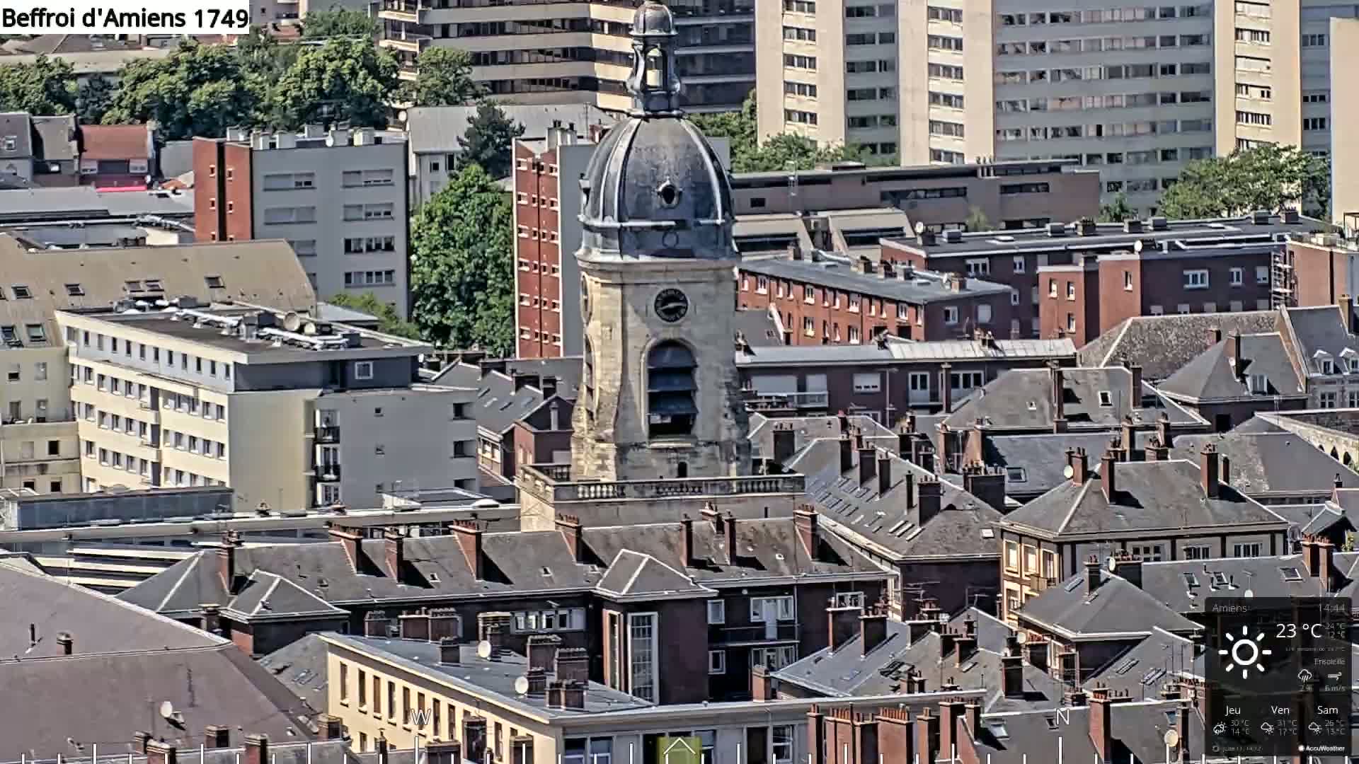 A high-angle, sunny view of a European city, featuring a domed clock tower rising above a dense collection of dark-roofed buildings.