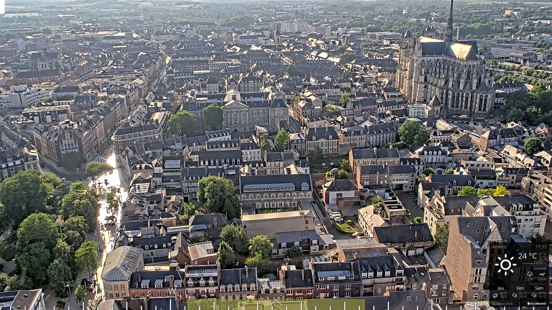 A high-angle, sunny view of a densely packed European city, featuring a large cathedral and a long street lined with trees.