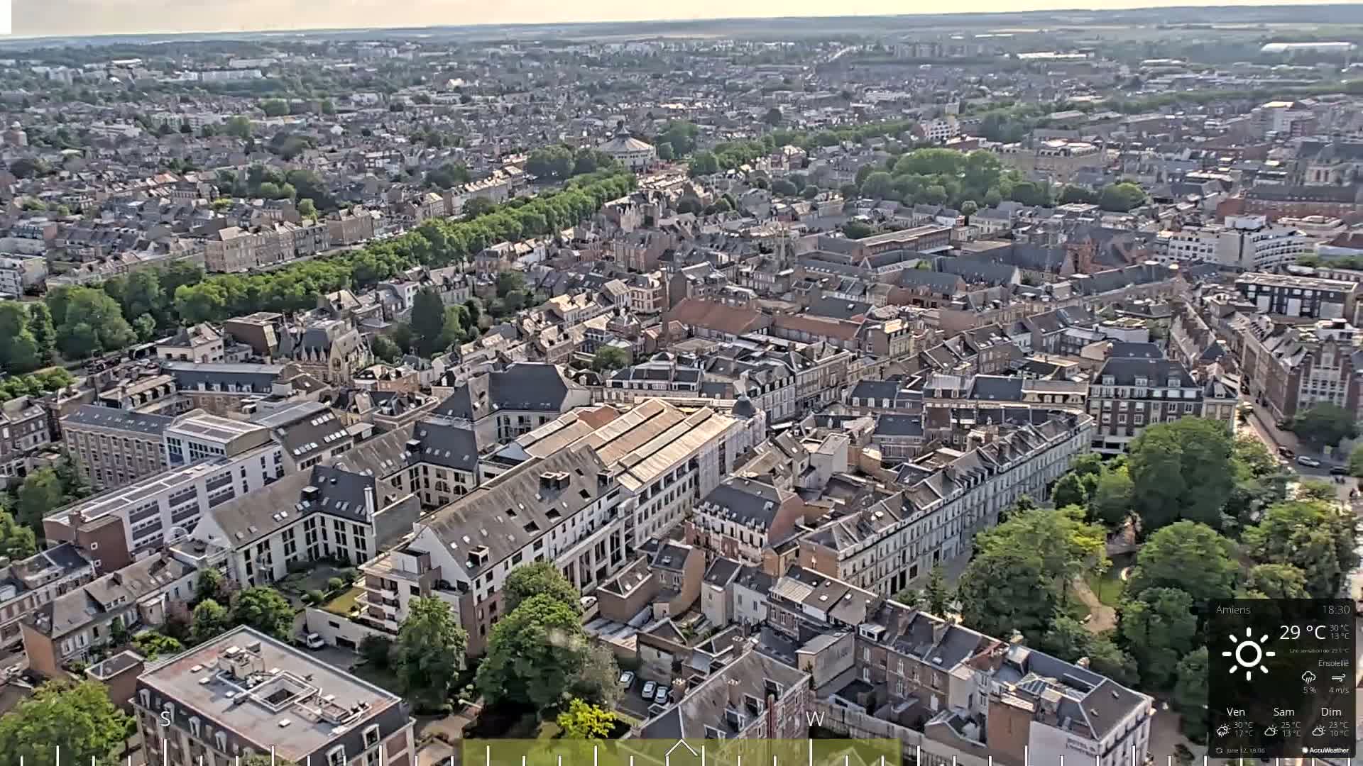 A high-angle, wide shot of a densely populated city on a sunny day shows many closely packed buildings with green trees and parks interspersed.