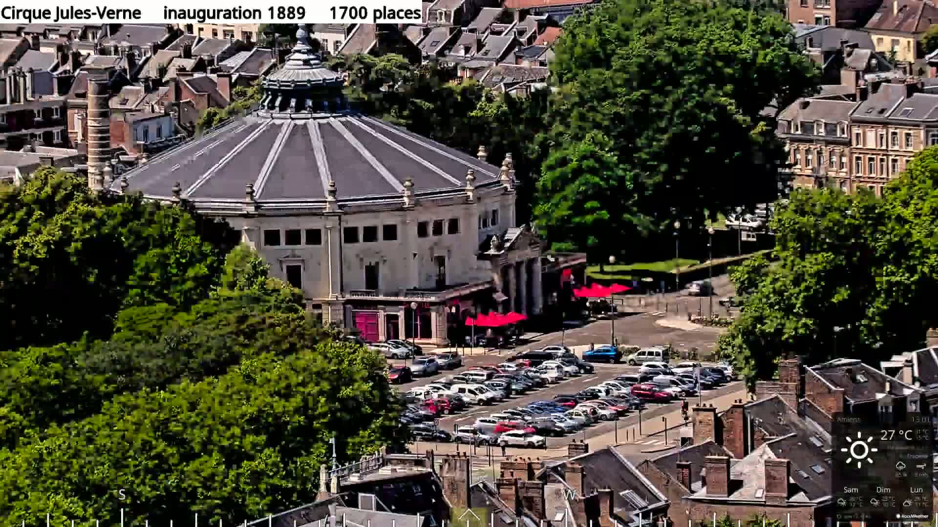 A large, round, light-colored building with a dark dome sits in a parking lot surrounded by lush green trees and other buildings on a sunny day.
