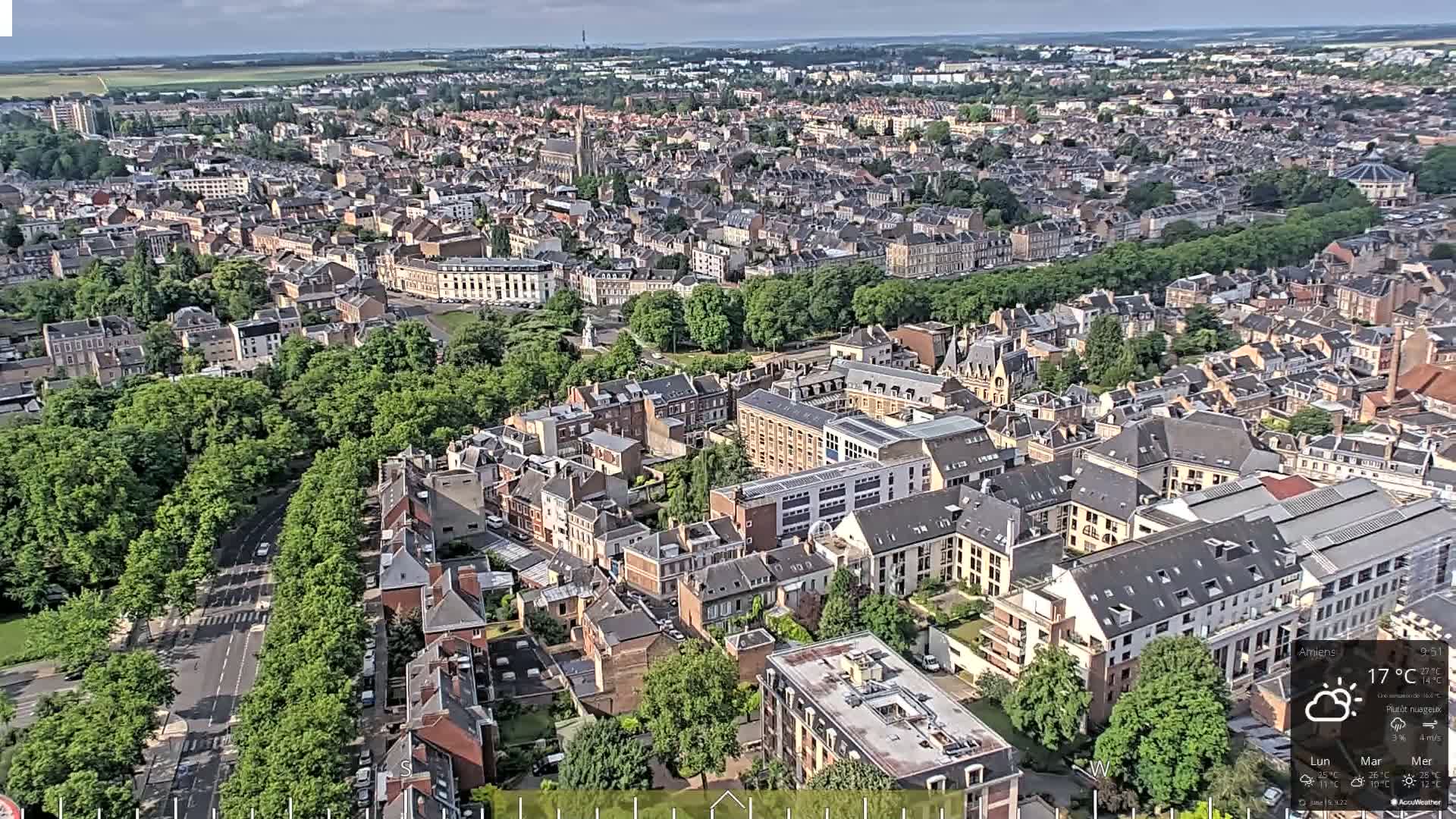 An aerial view shows a densely populated city with many buildings and tree-lined streets under partly cloudy skies.
