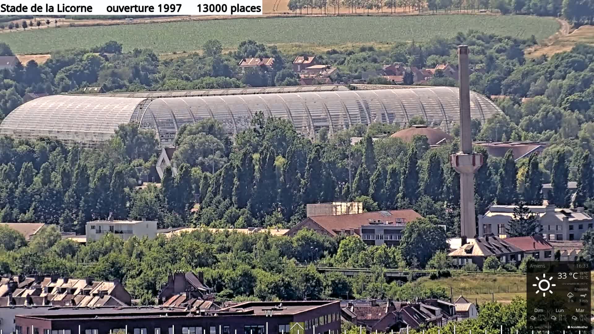 A large, curved, glass-roofed stadium sits among green trees and residential buildings under a sunny sky.