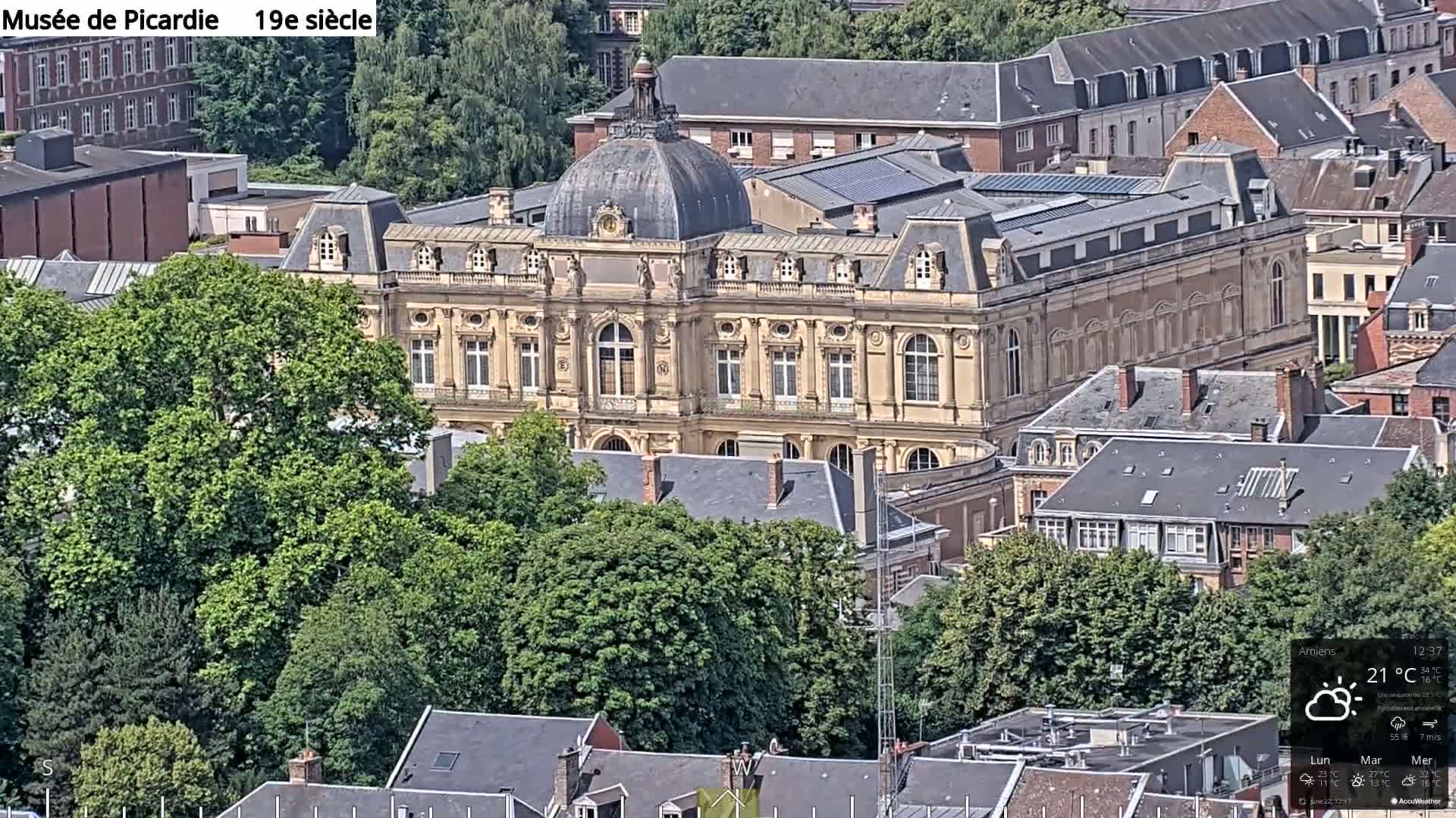 An elevated view of a large, light-beige building with a dome, surrounded by other buildings and lush green trees, on a partly cloudy day.