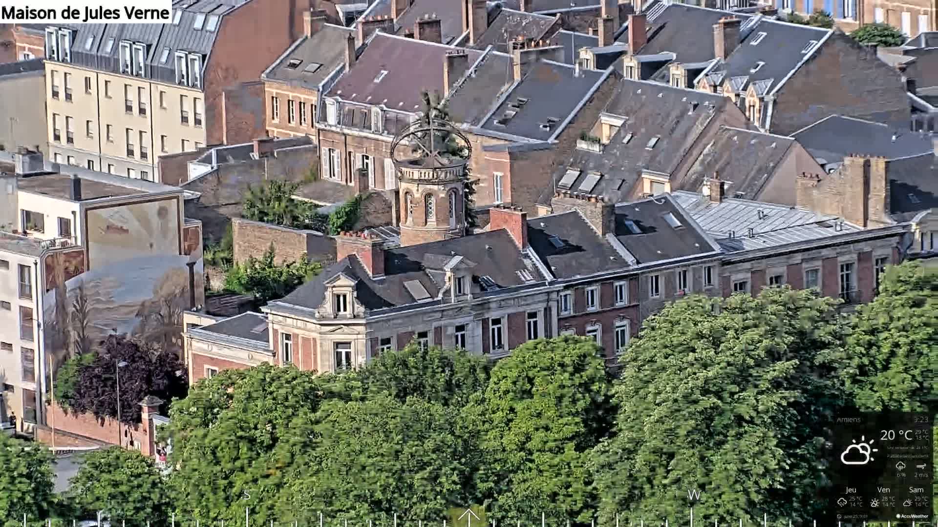 An aerial view shows a cluster of brick buildings, one with a tower topped by a weathered-looking sphere, partially obscured by lush green trees under a partly sunny sky.