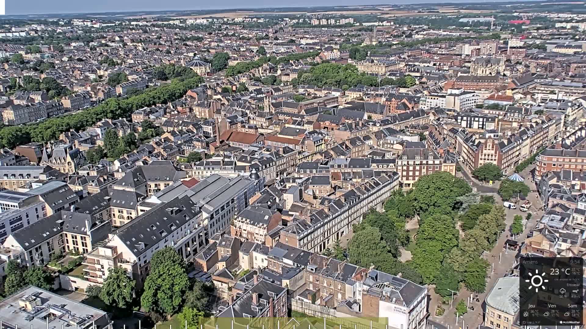 An aerial view of a densely populated city on a sunny day, showing a mix of residential and commercial buildings with green spaces interspersed.
