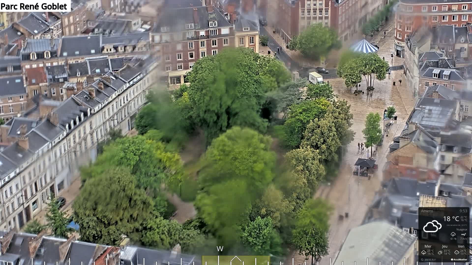 A flooded city square surrounded by buildings and a park is seen from an aerial view on a cloudy day.