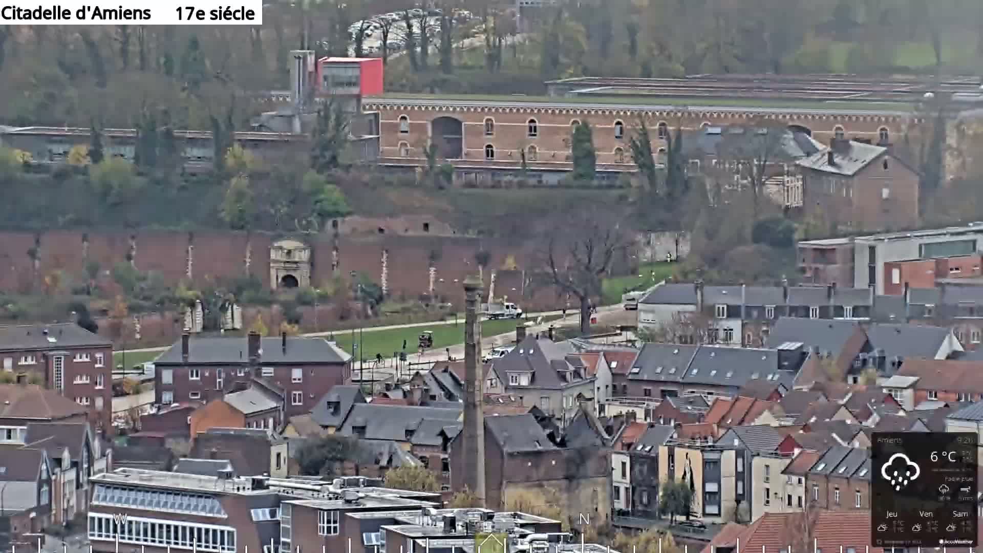 An elevated, wide-angle view captures a sprawling European townscape featuring dense clusters of buildings with varied roofs, a prominent tall brick chimney, and a large ancient stone wall with an arched gateway in the mid-distance, all under an overcast sky typical of a cool, possibly autumnal day.