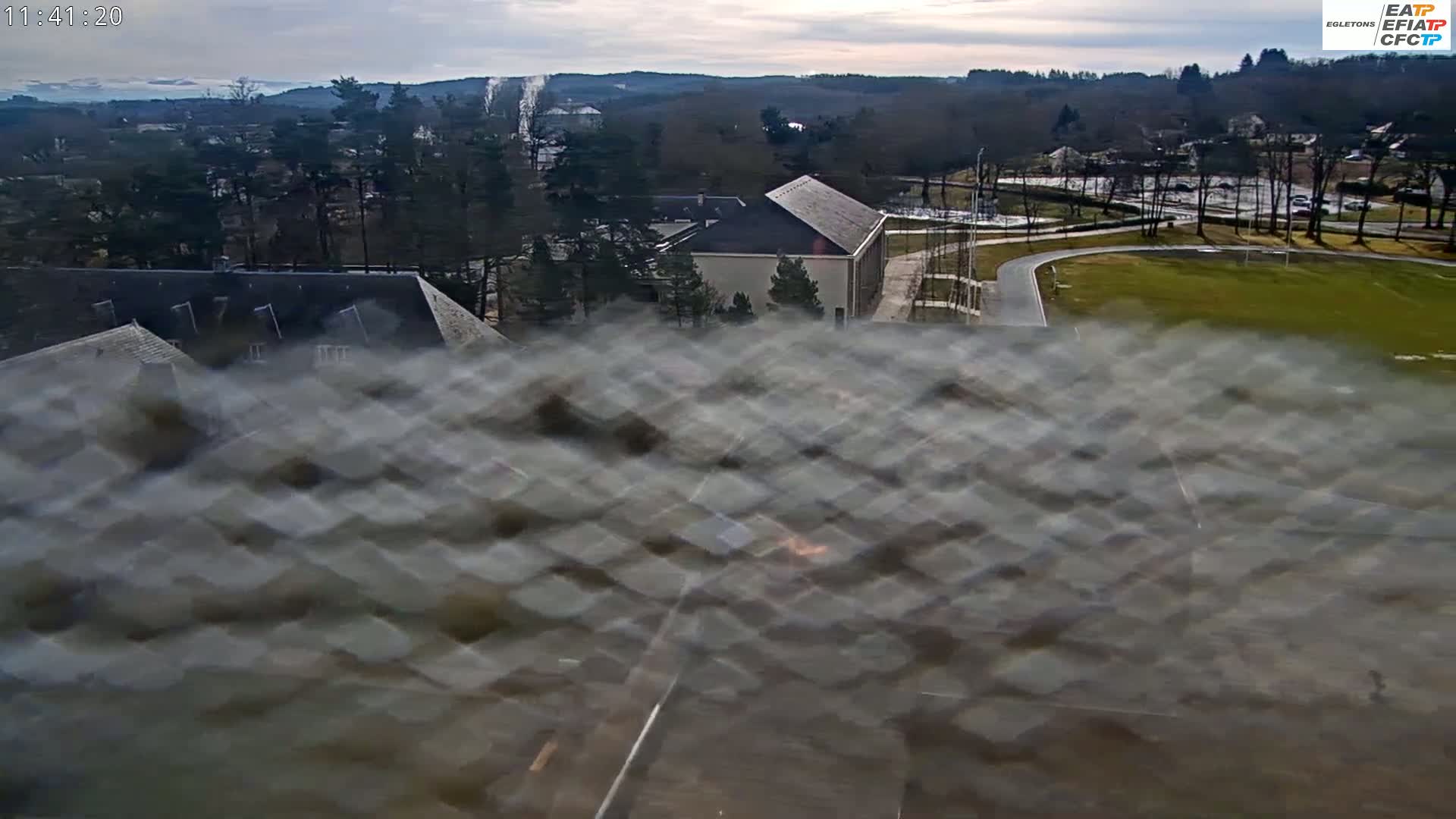 A mostly cloudy day reveals a view of buildings and a large grassy field, seen through a blurry window.
