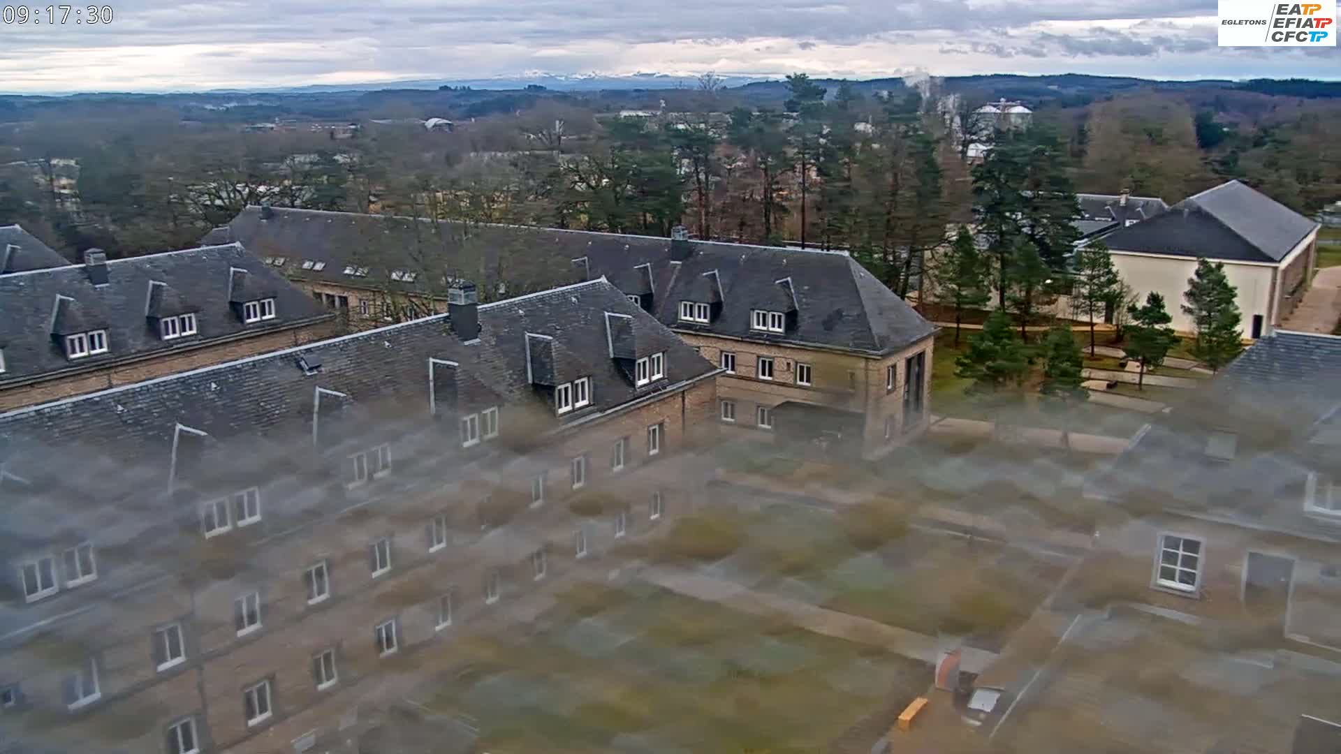 An aerial view of a complex of low-rise buildings with slate roofs, surrounded by trees under an overcast sky.