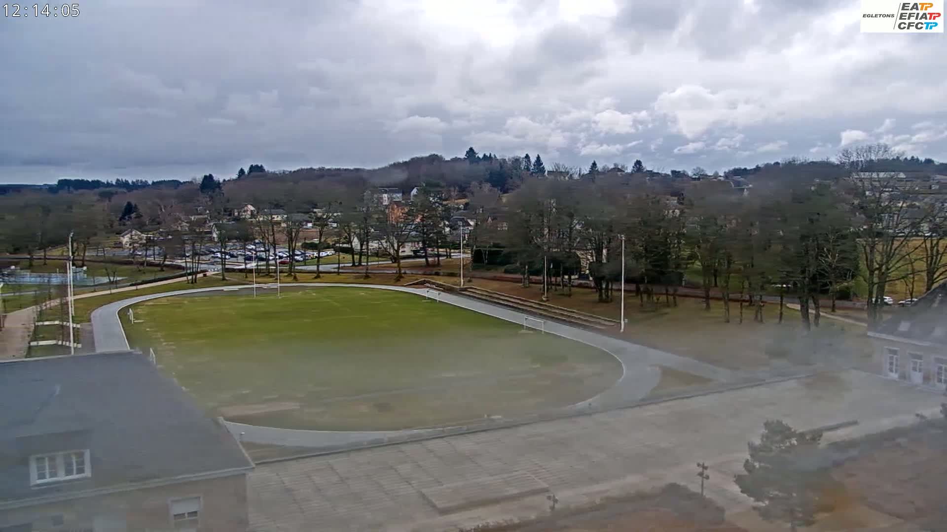 A high-angle view shows a running track surrounding a grassy field, situated next to a town under a cloudy sky.