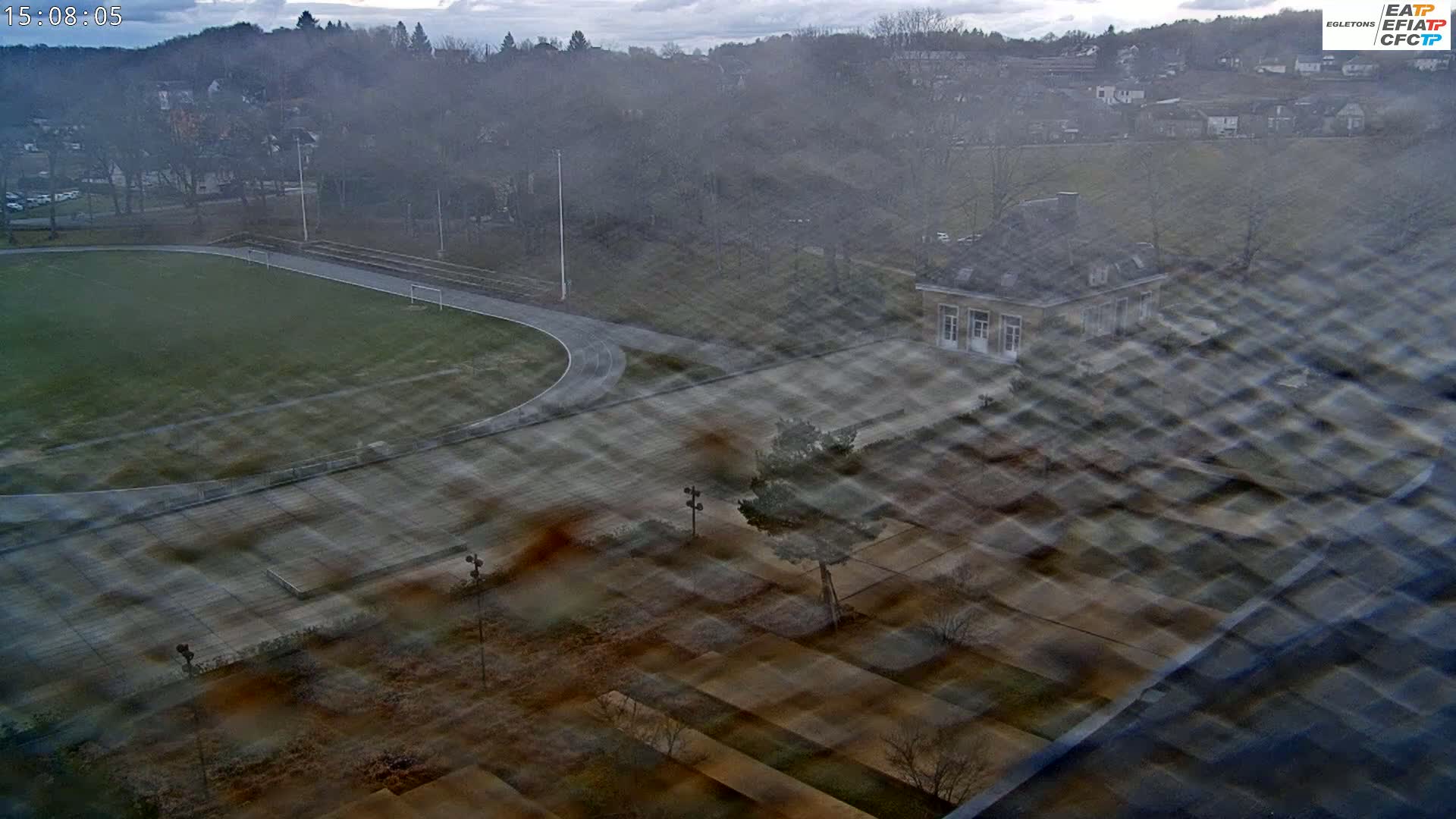 An overcast day reveals a high-angle view of a running track, a small building, and a surrounding landscape with trees and houses in the distance.