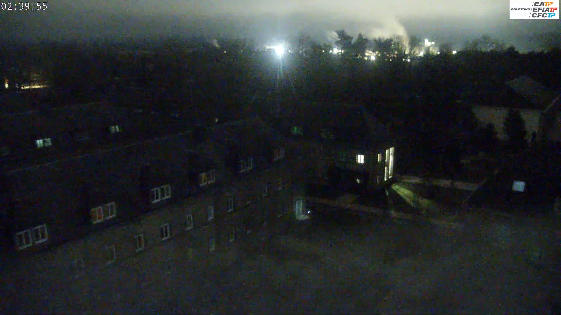A nighttime aerial view shows several dark buildings with dimly lit windows under a cloudy sky.