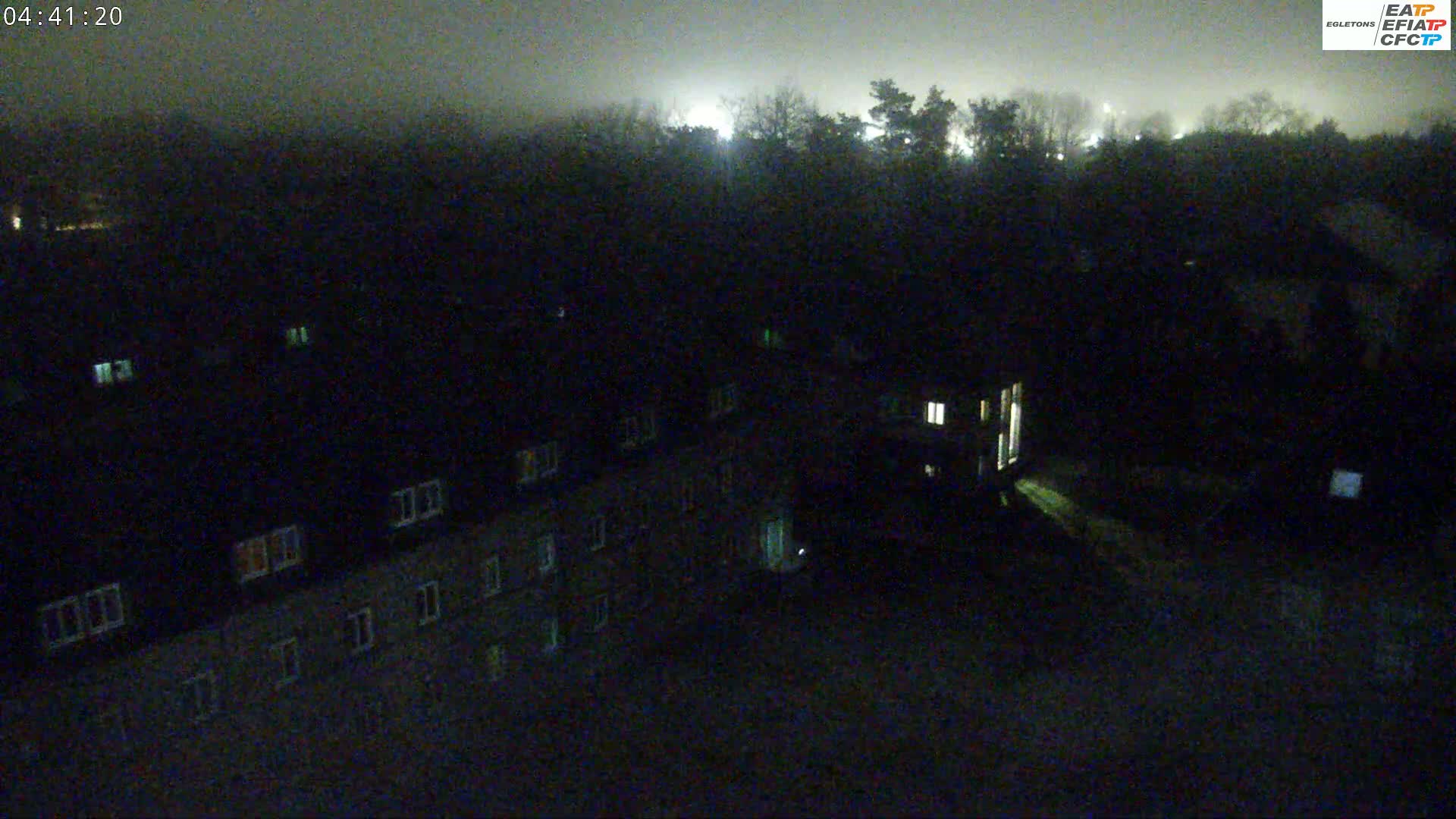 A nighttime, foggy view shows a dark building with illuminated windows, and a dark line of trees in the background.
