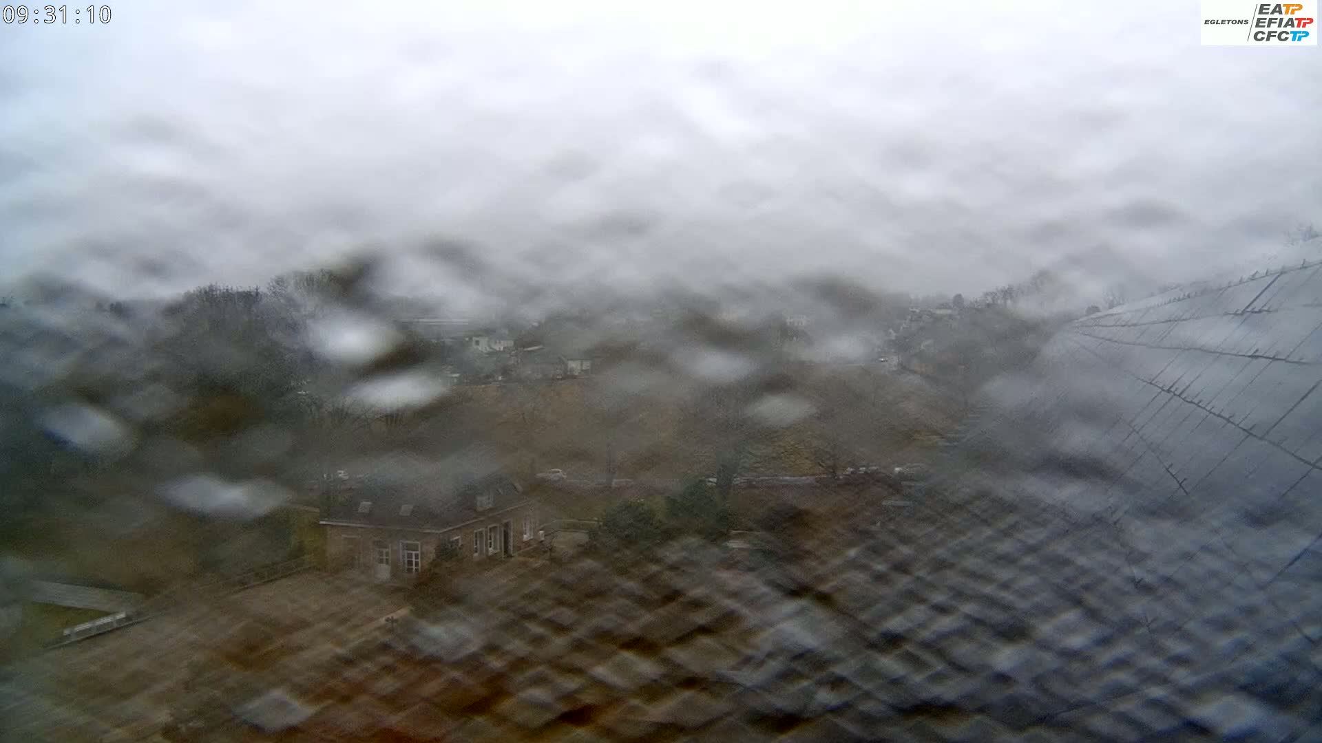 A rain-streaked view shows a blurry landscape of houses and trees under a gray, overcast sky.