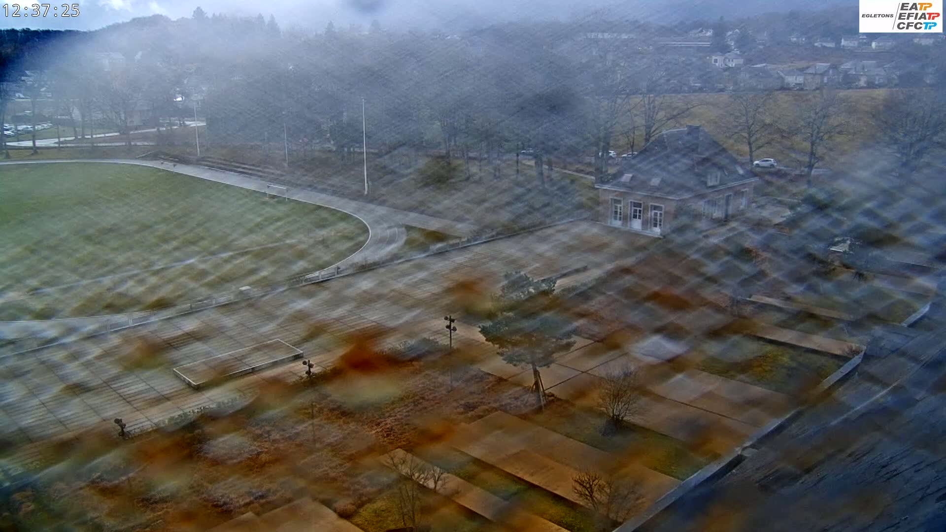 A high-angle view of a sports field and surrounding grounds on a misty day, showing a small stone building and landscaping features.