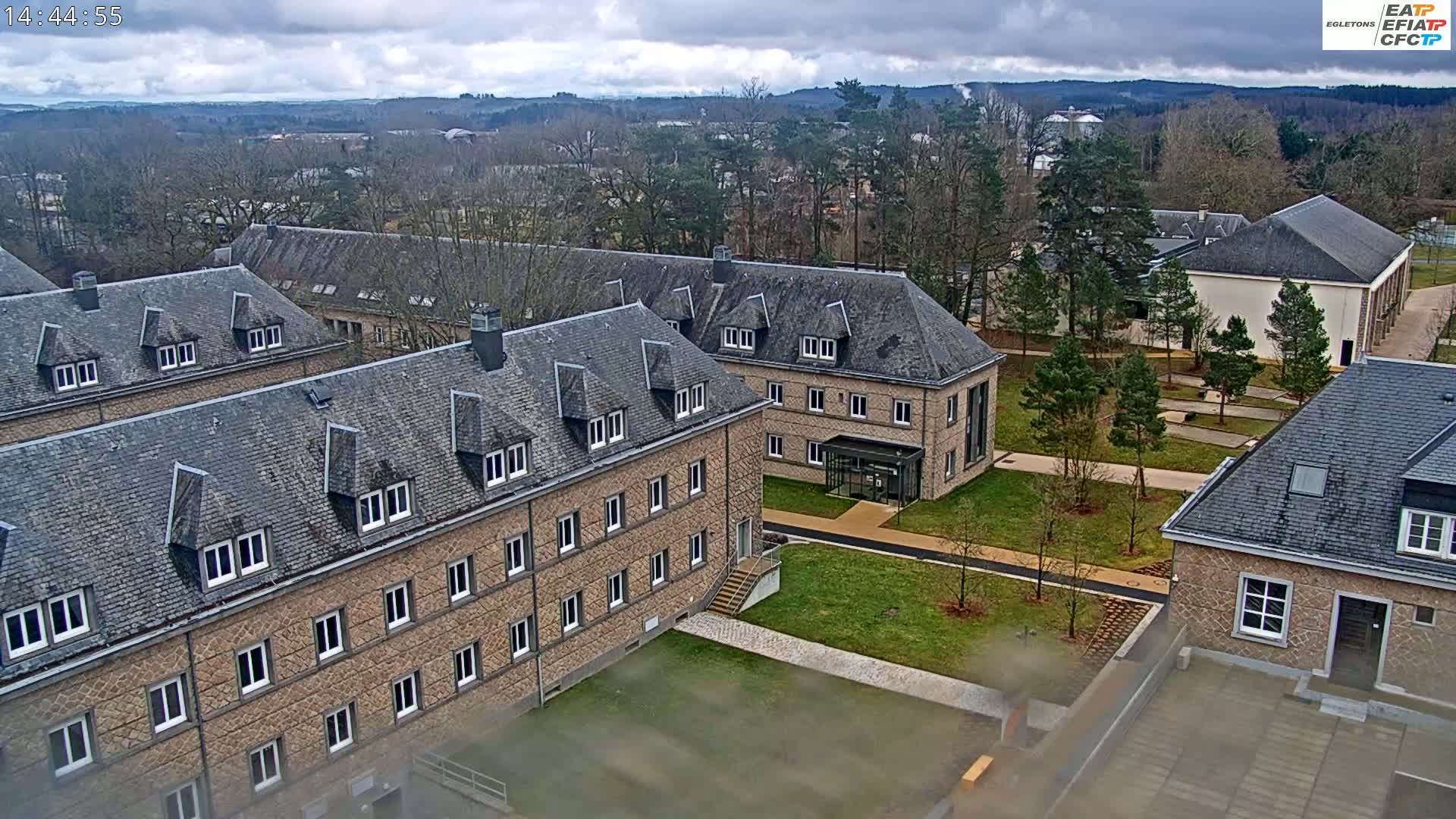A complex of low-rise stone buildings with slate roofs sits in a landscaped courtyard on an overcast day, surrounded by bare trees and distant hills.