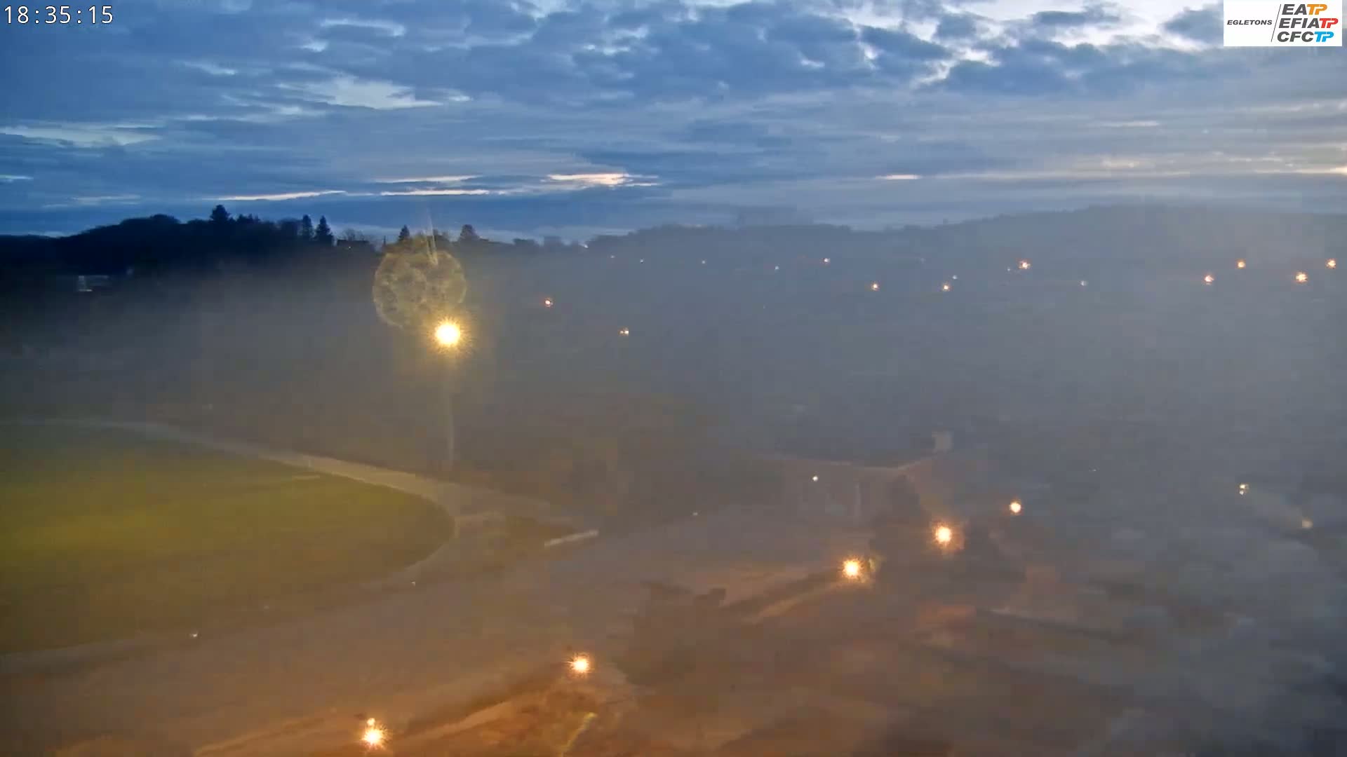 A hazy, twilight view shows a wide expanse of land with scattered lights, including a field, a road, and distant buildings under a cloudy sky.