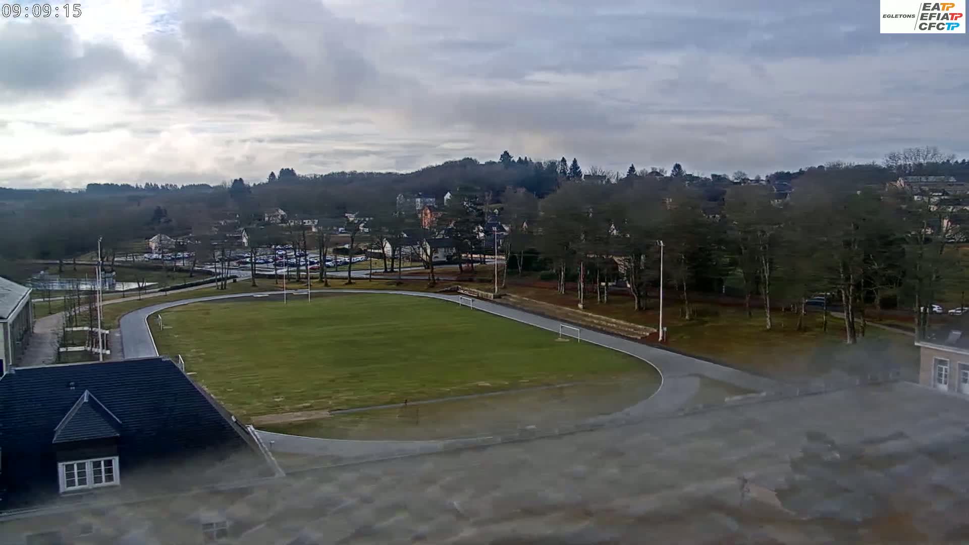 An overcast day reveals a running track and soccer field nestled beside a small town, surrounded by trees and buildings.