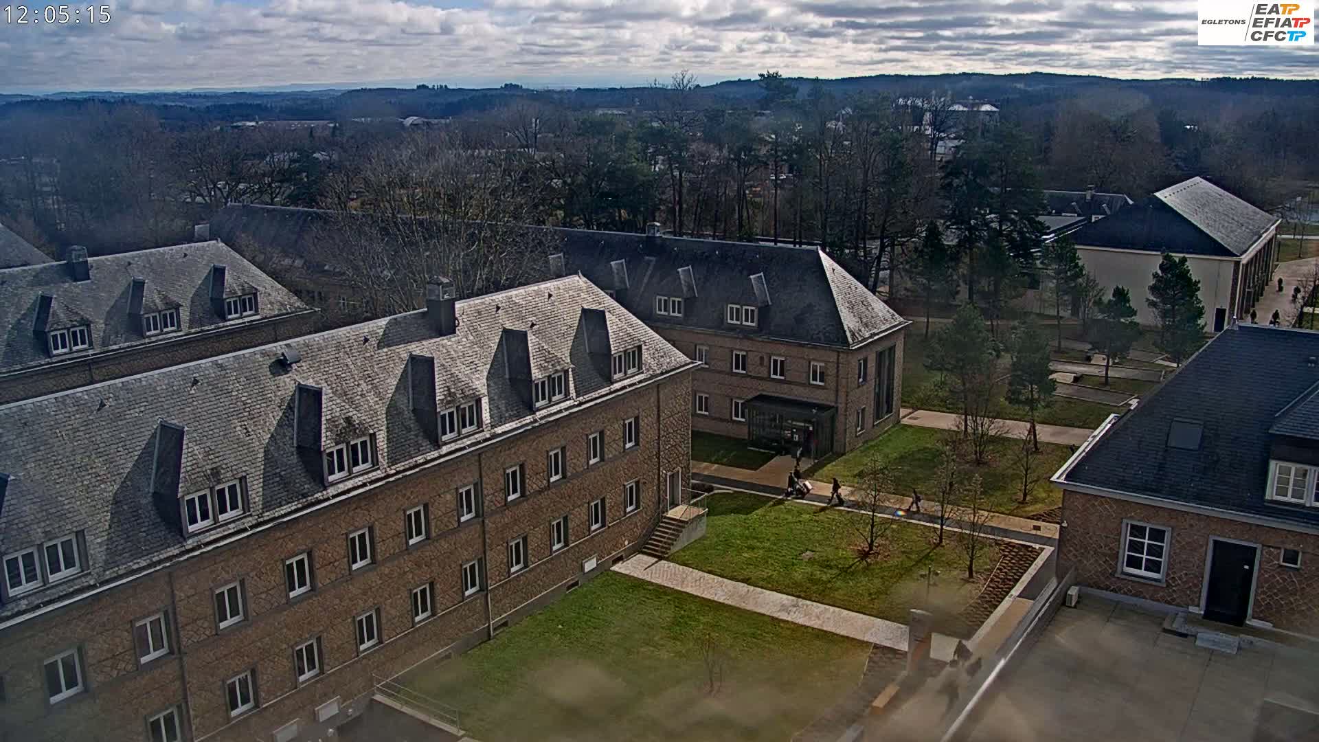 A high-angle view of a campus with several brick buildings under a partly cloudy sky, showing a grassy courtyard with a few people walking.