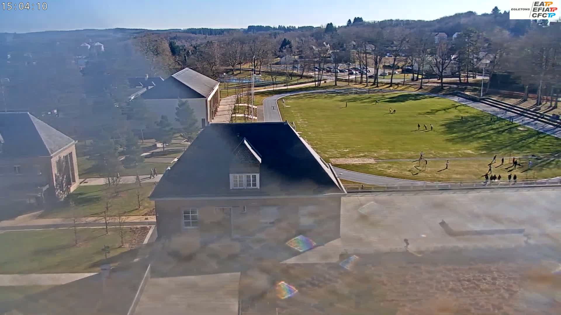 A high-angle, hazy view shows a school campus on a sunny day with people using a grassy athletic field and track.