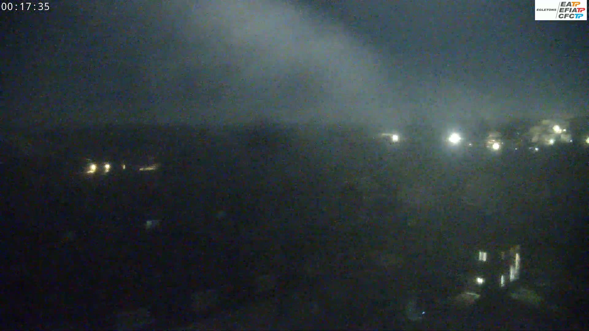 A nighttime, hazy view shows a dark landscape with scattered lights from buildings and vehicles under an overcast sky.