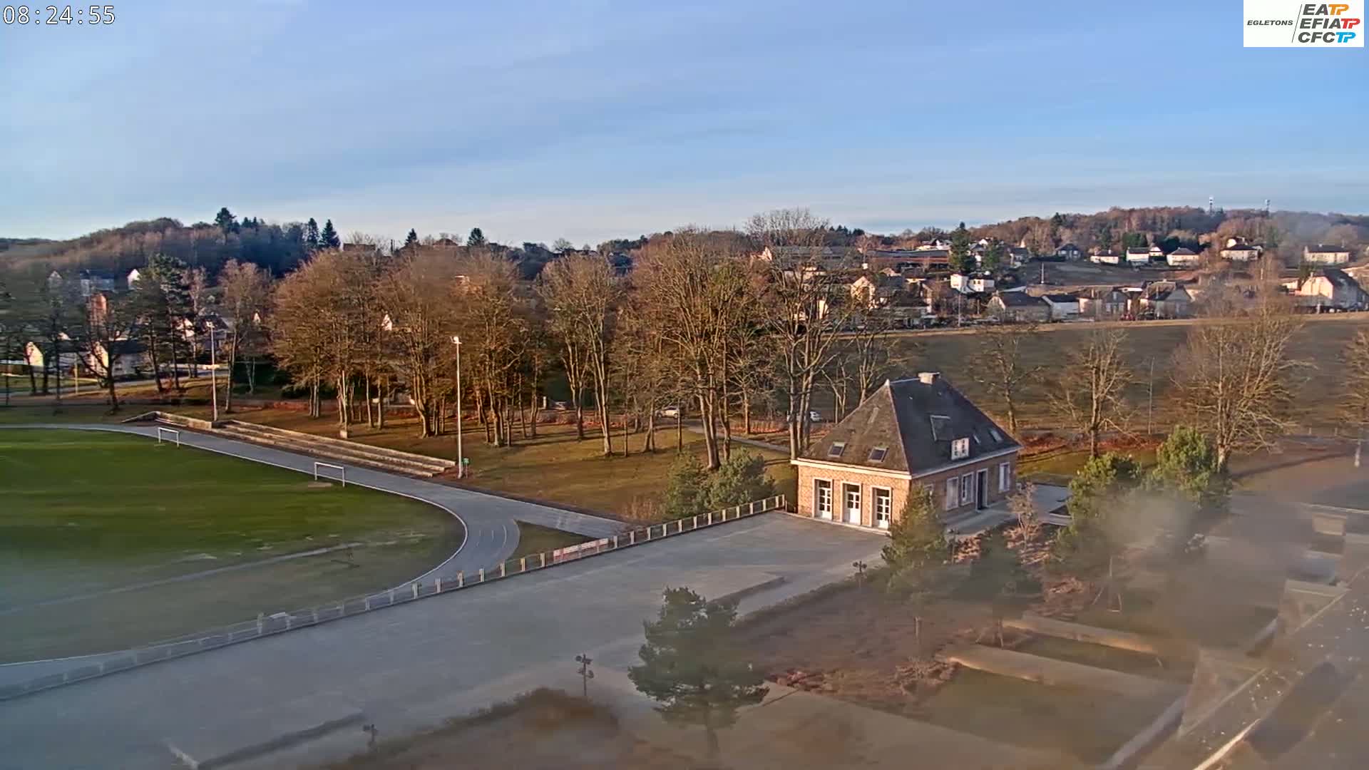 An aerial view shows a running track and a small brick building next to a park under a clear, sunny sky.