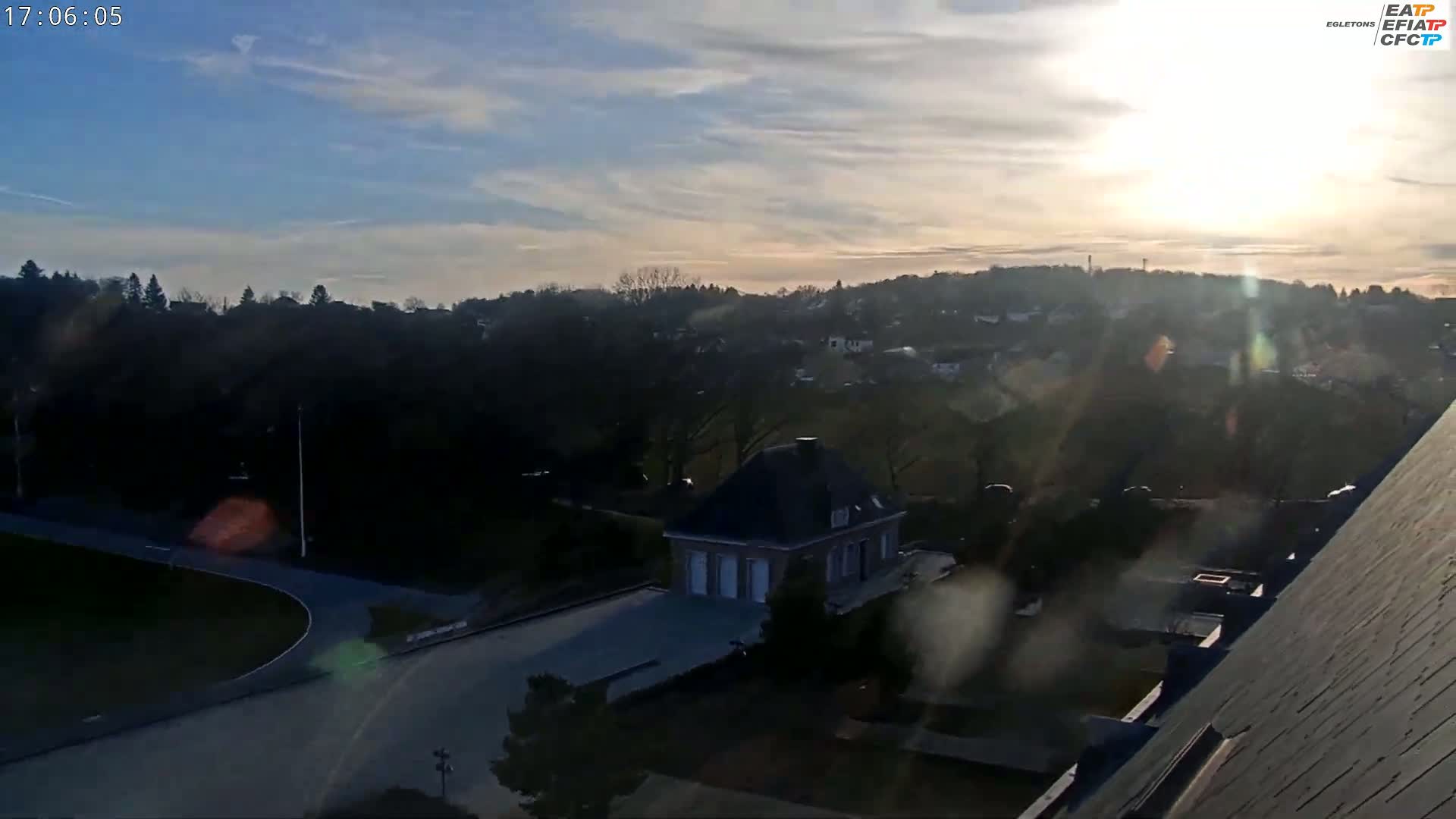 A high-angle, long shot shows a partly sunny, hazy day overlooking a residential area with a small building in the foreground, and a road curving through a park-like setting.