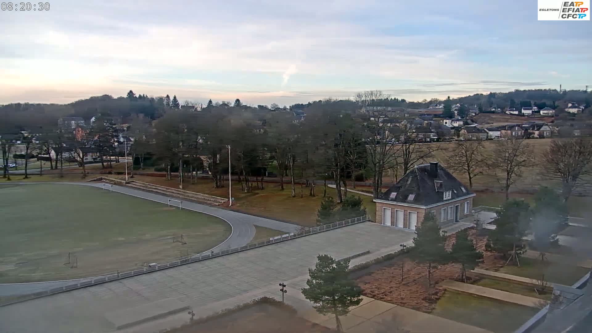 A high-angle view of a running track and small building, situated in a park-like setting next to a small town under a mostly clear sky.