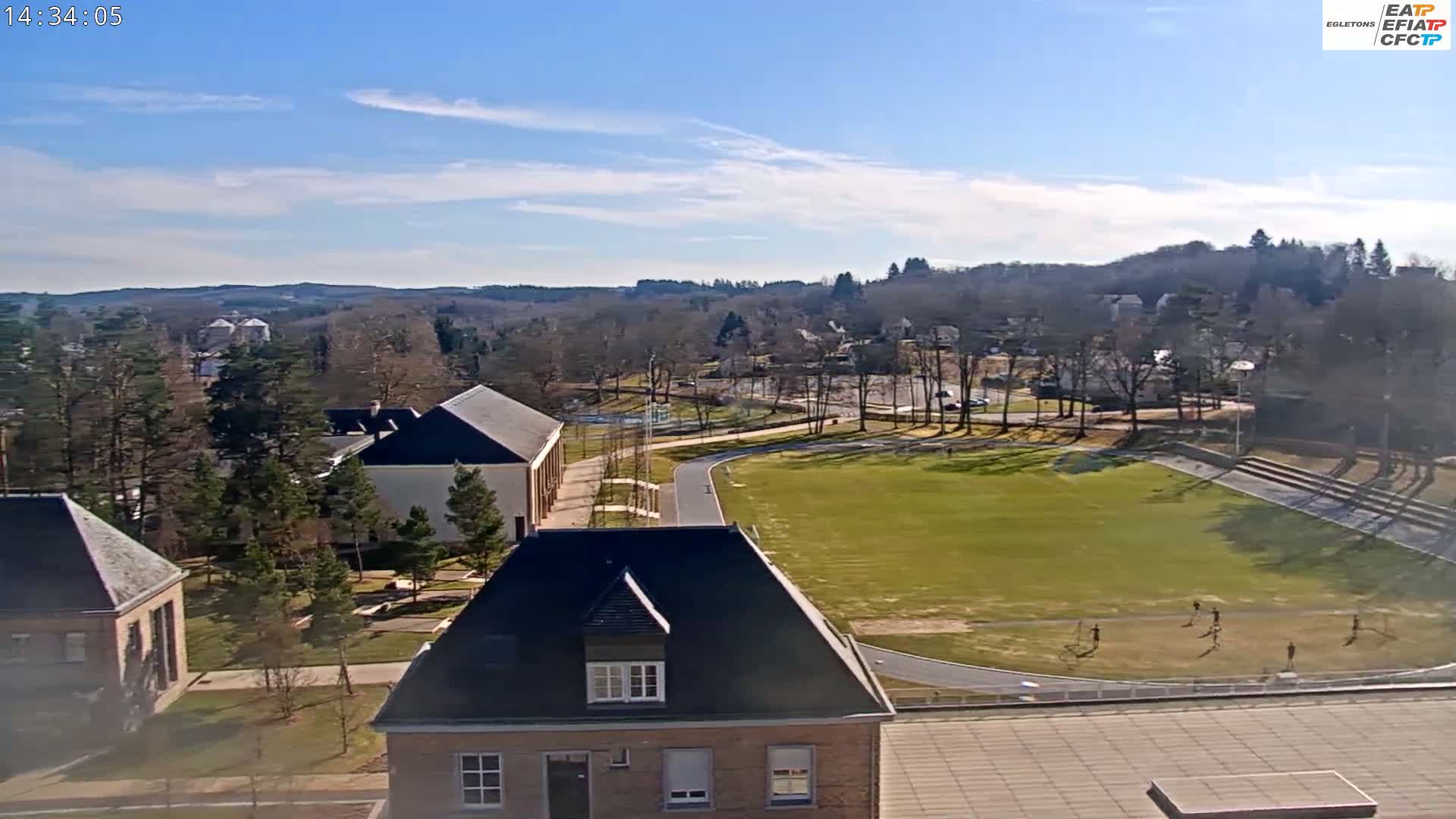 A high-angle view shows a campus on a sunny day with a large grassy field, several buildings, and a few people on the field.
